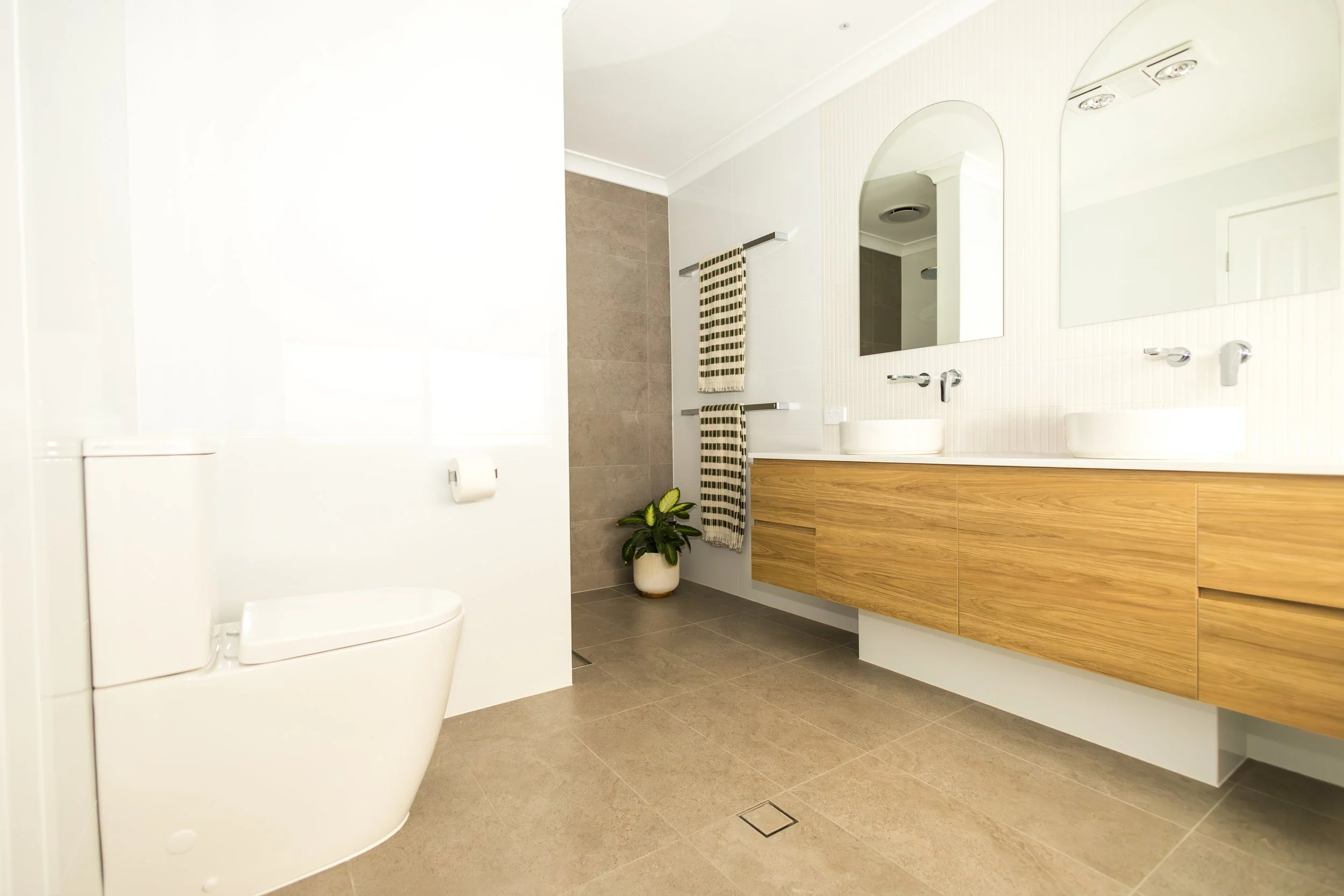 Modern bathroom with dual sinks, large mirrors, wooden vanity, tiled walls and floors, a potted plant, and hanging towels.