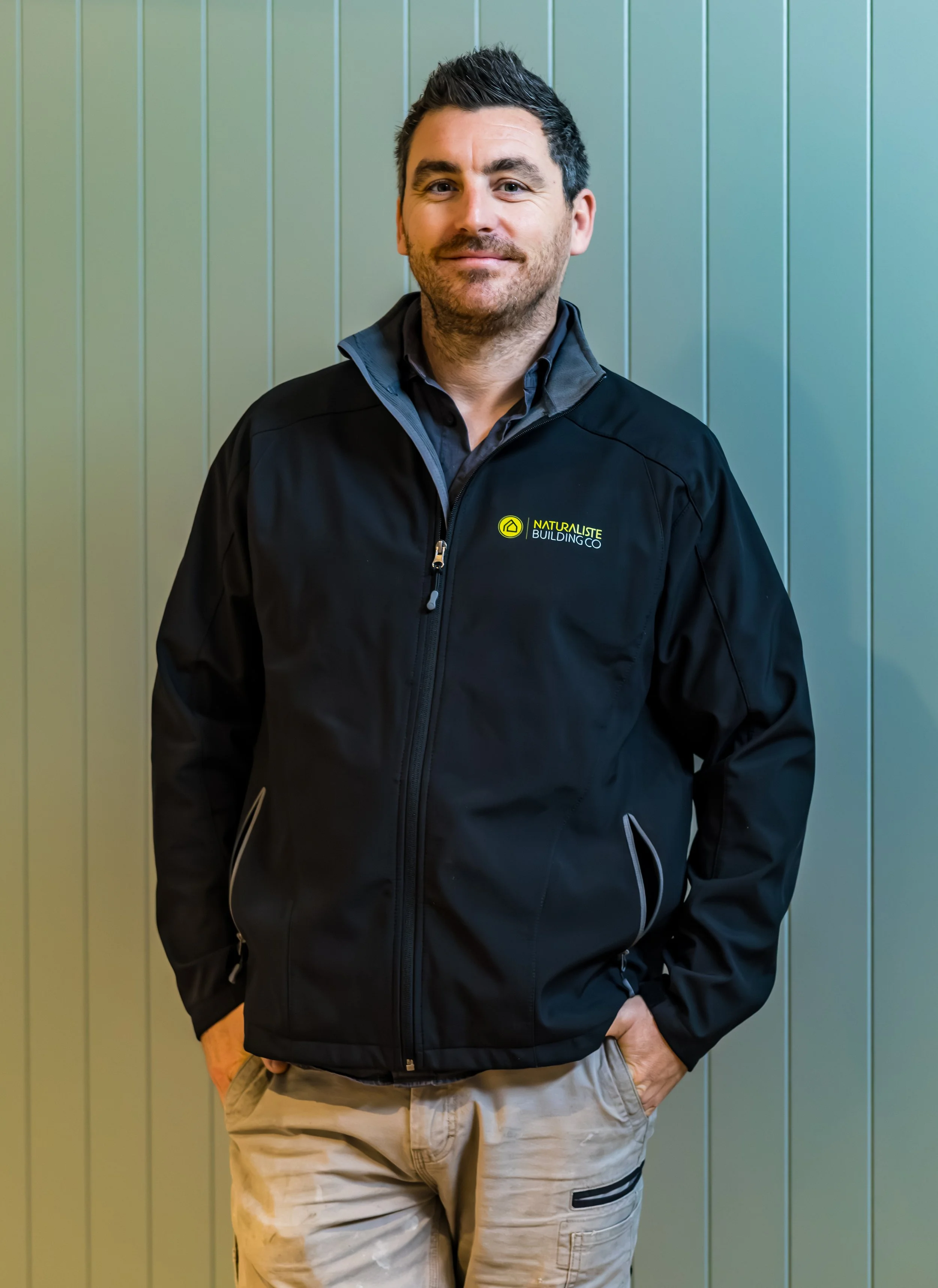 A man wearing a black jacket with the logo and text 'NATURALISTE BUILDING CO' standing in front of a light green paneled wall, with his hands in his pockets, looking at the camera and smiling.