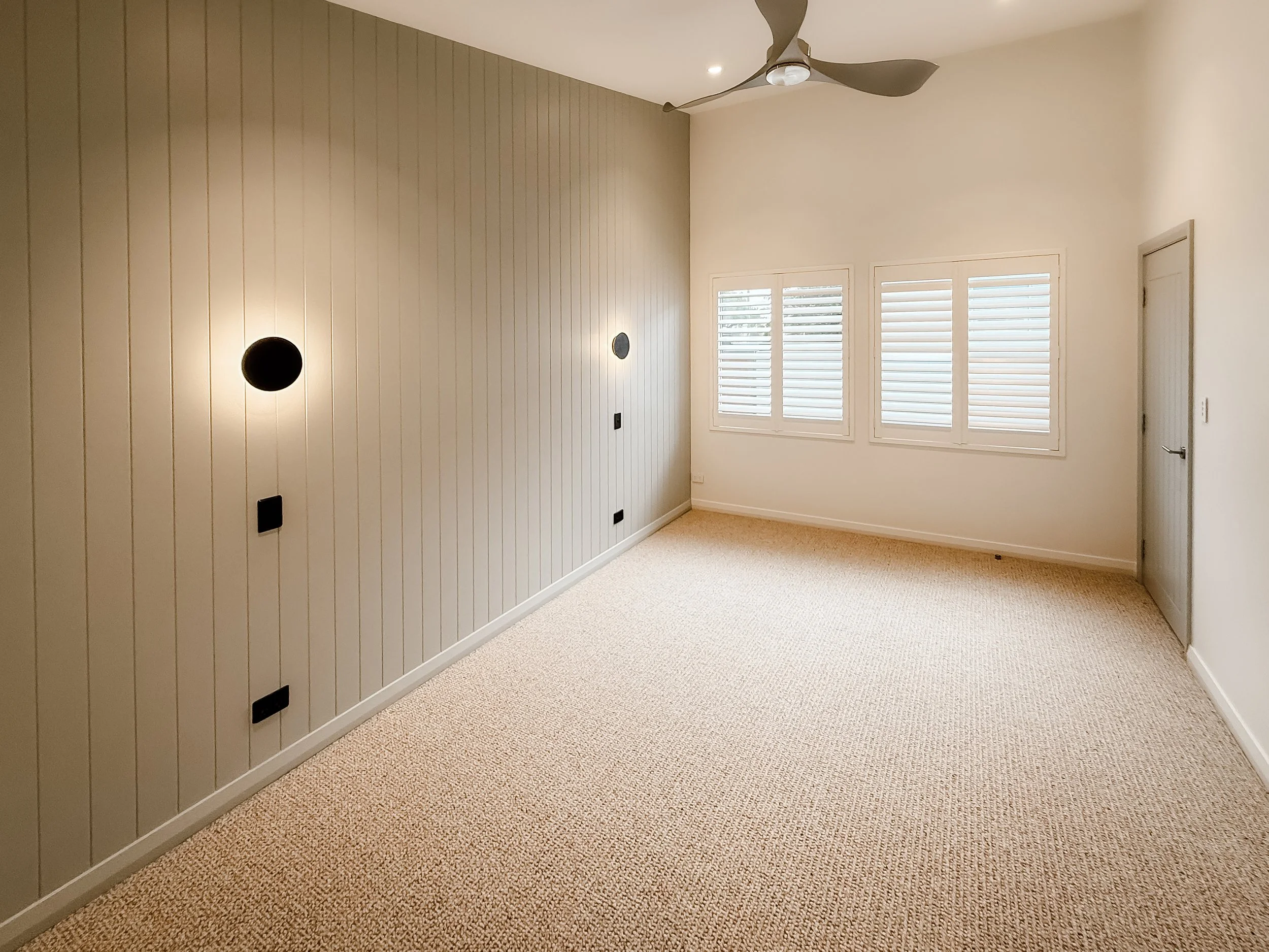 Empty room with beige carpet, white walls, three windows with white shutters, a door with a silver handle, and a ceiling fan. One wall has vertical beadboard paneling and installed black wall fixtures.