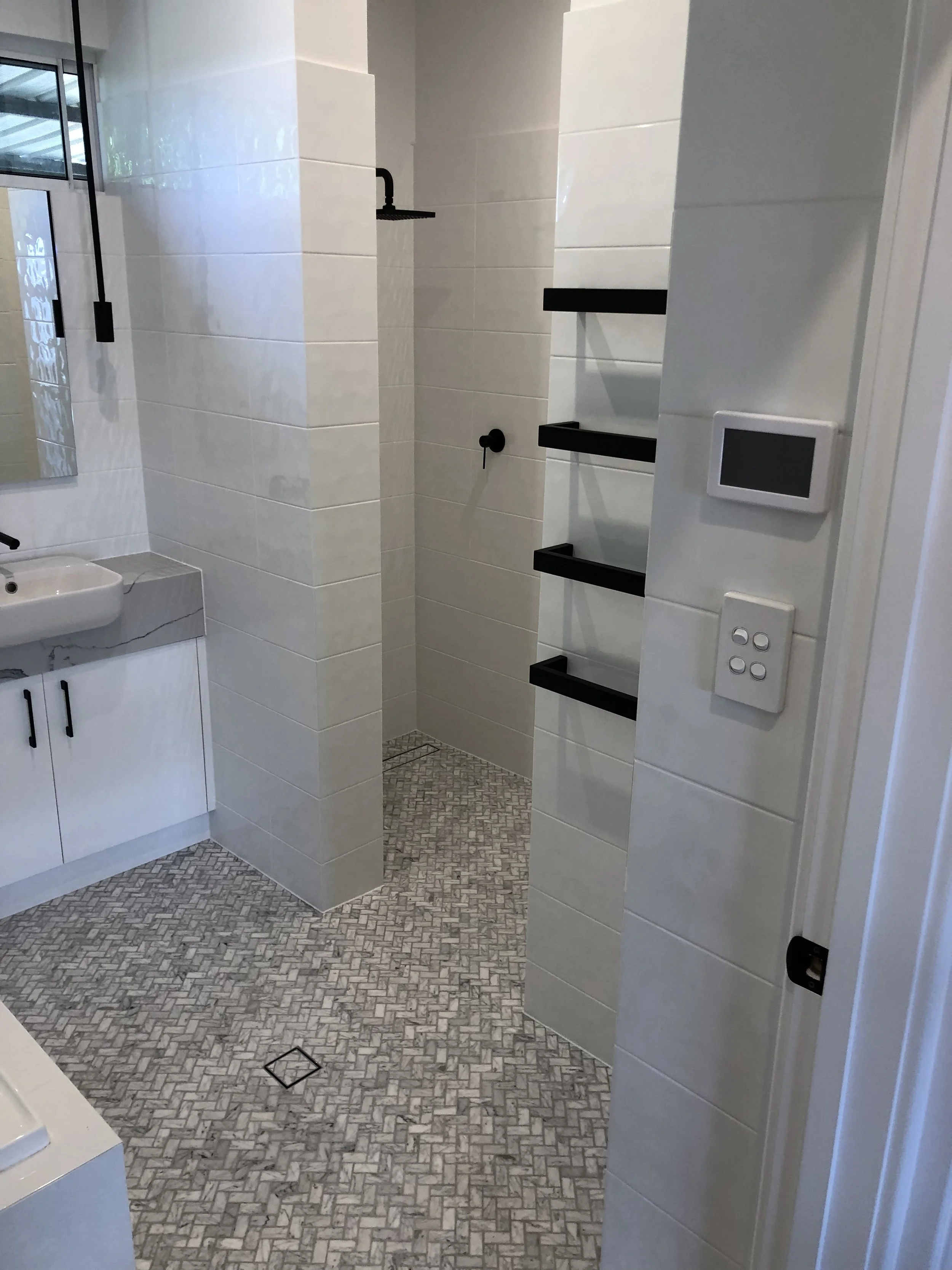 Bathroom with white tile walls, black shelves, small sink, and a walk-in shower with dark fixtures.