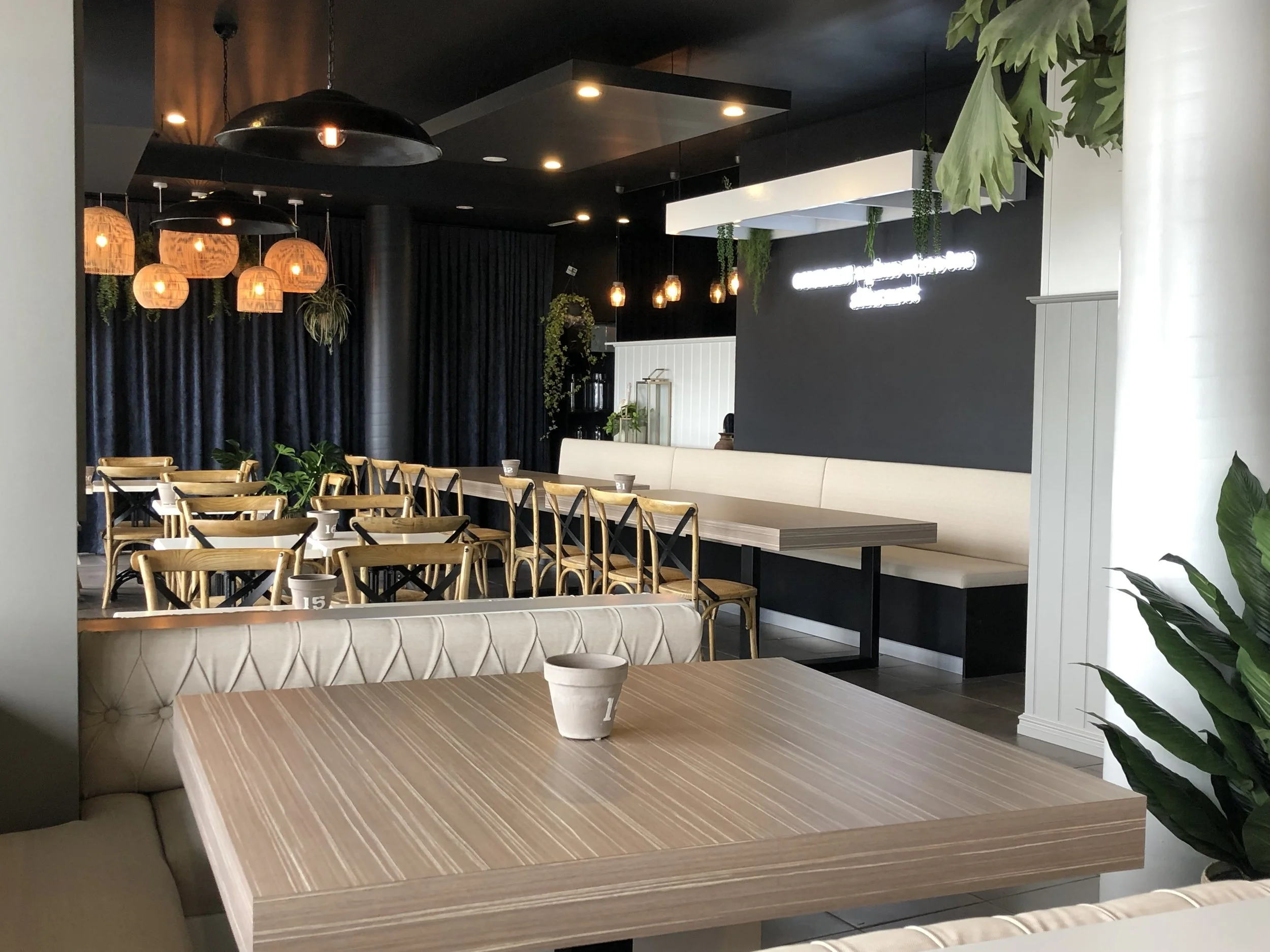 Empty restaurant seating area with a wooden table in the foreground, surrounded by beige and wooden chairs, and a row of white cushioned booth seating along the wall in the background. Hanging lights and green plants decorate the space, with dark cur