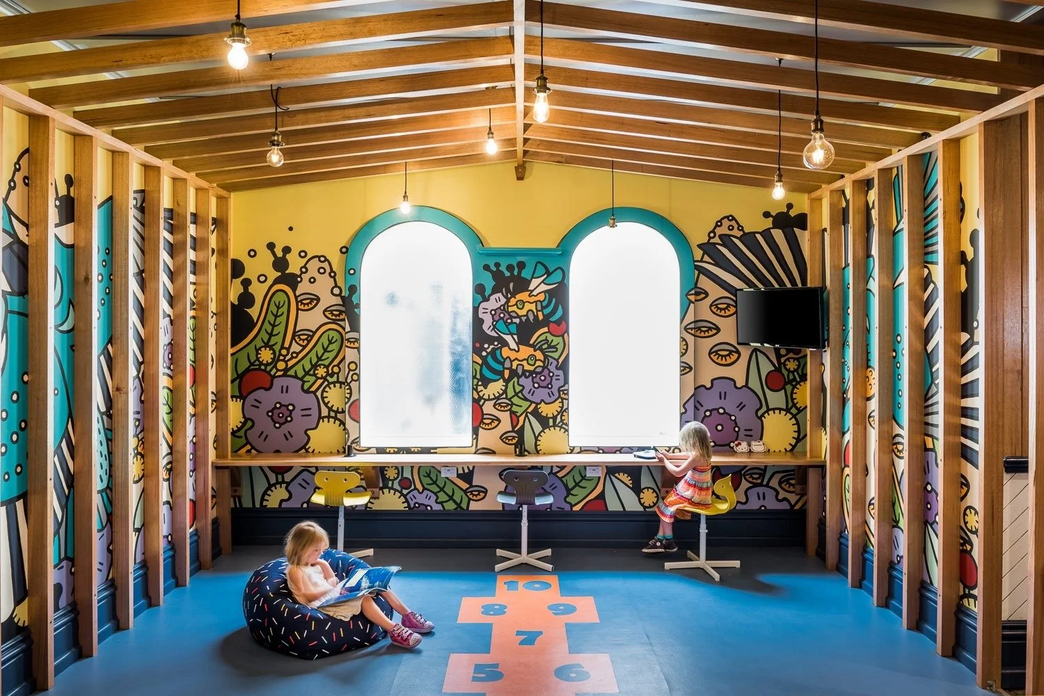 commercial build of a Childrens play room playing in a colorful, mural-decorated indoor playroom with a hopscotch on the floor, wooden beams, hanging light bulbs, and a small TV mounted on the wall. 