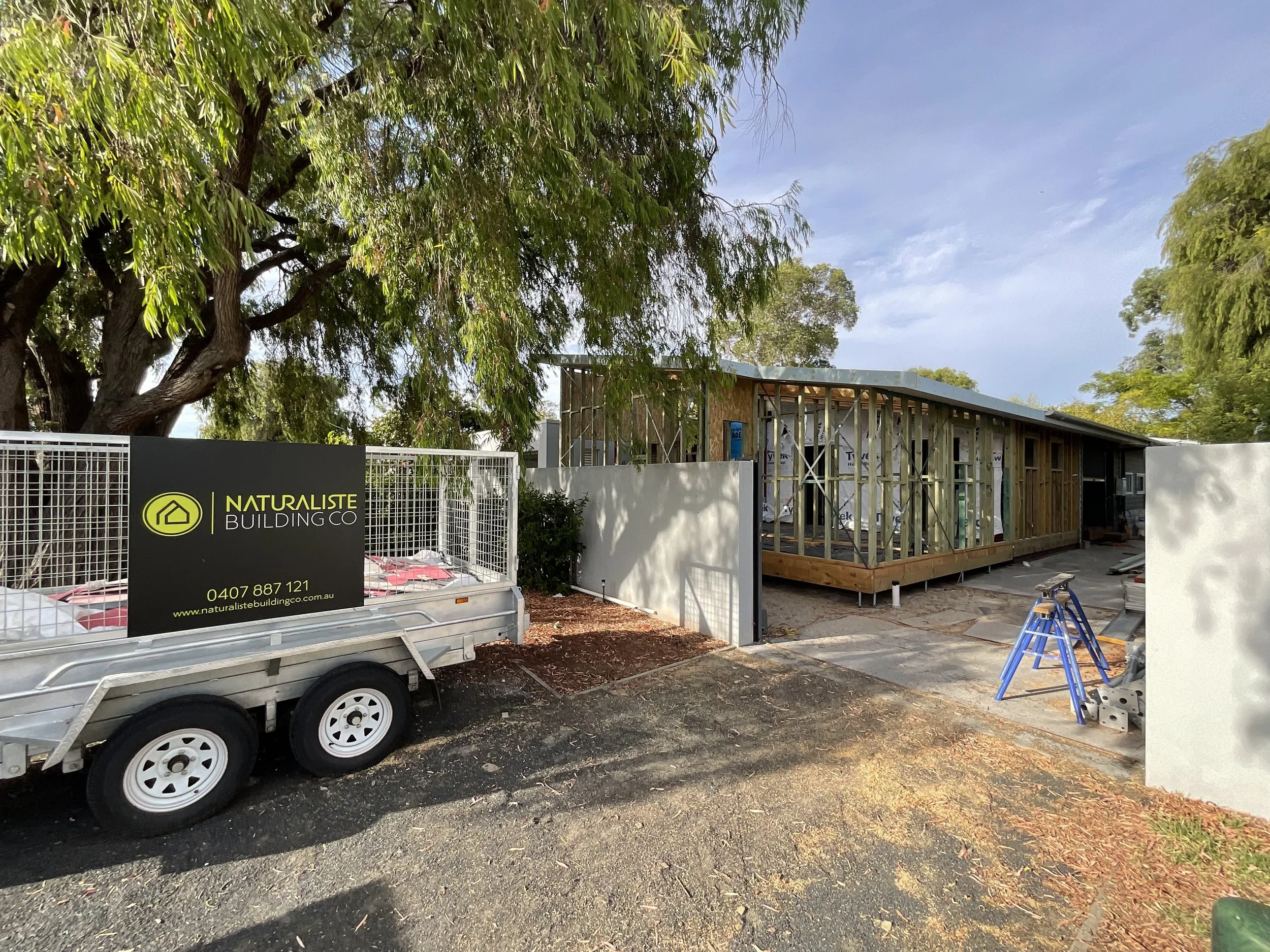 A house under construction with exposed wooden framing, situated behind white walls. There is a large tree to the left and a construction trailer with company branding for Naturaltiste Building Co. parked in front. Construction tools and materials ar