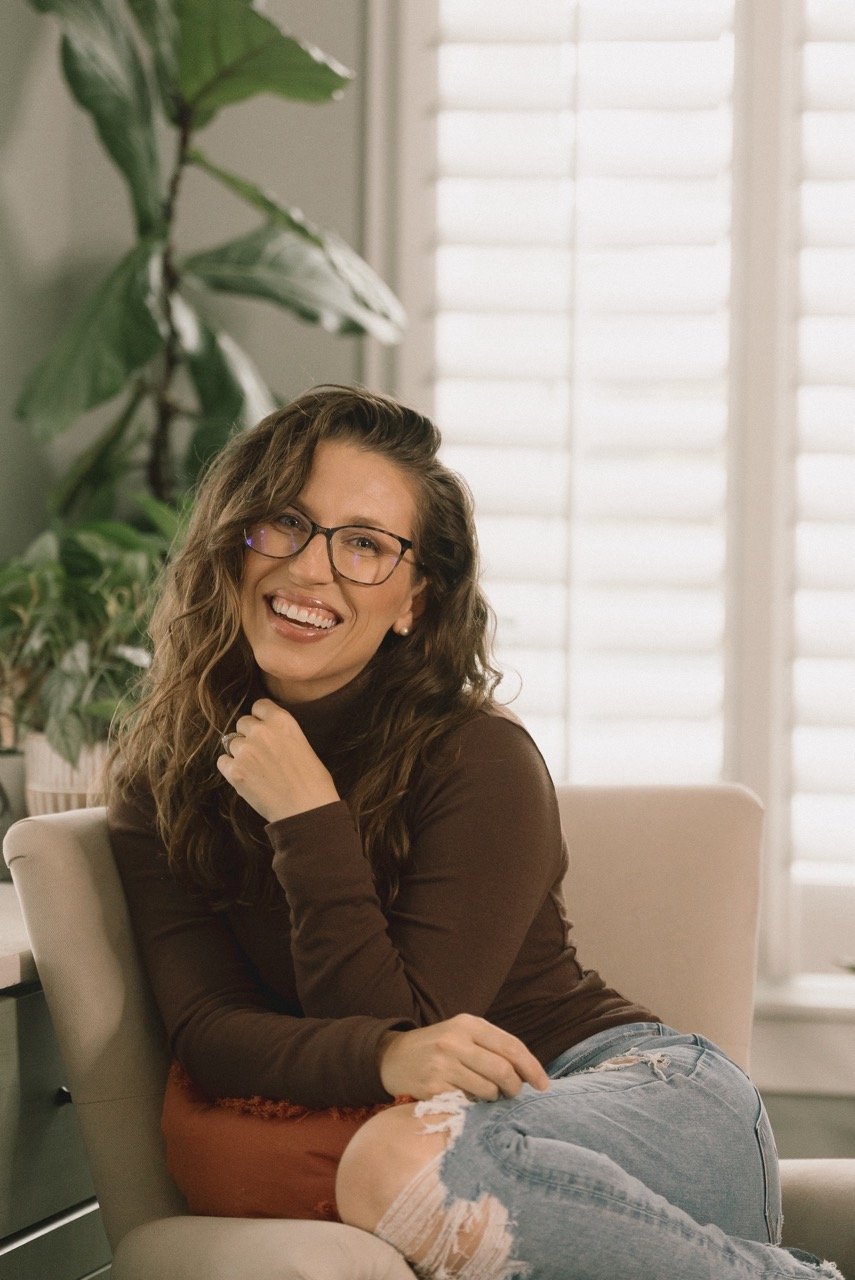 A woman with wavy brown hair and glasses, wearing a brown long-sleeve shirt and ripped jeans, smiling and sitting in a bright room with large windows and green plants.