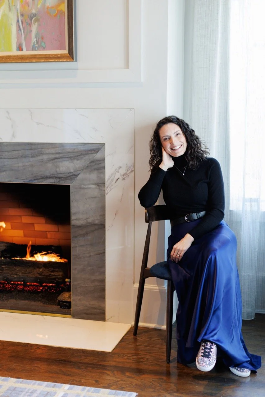 A woman with curly dark hair, wearing a black turtleneck sweater, blue satin skirt, and glittery sneakers, sitting on a black chair next to a fireplace with a smile.