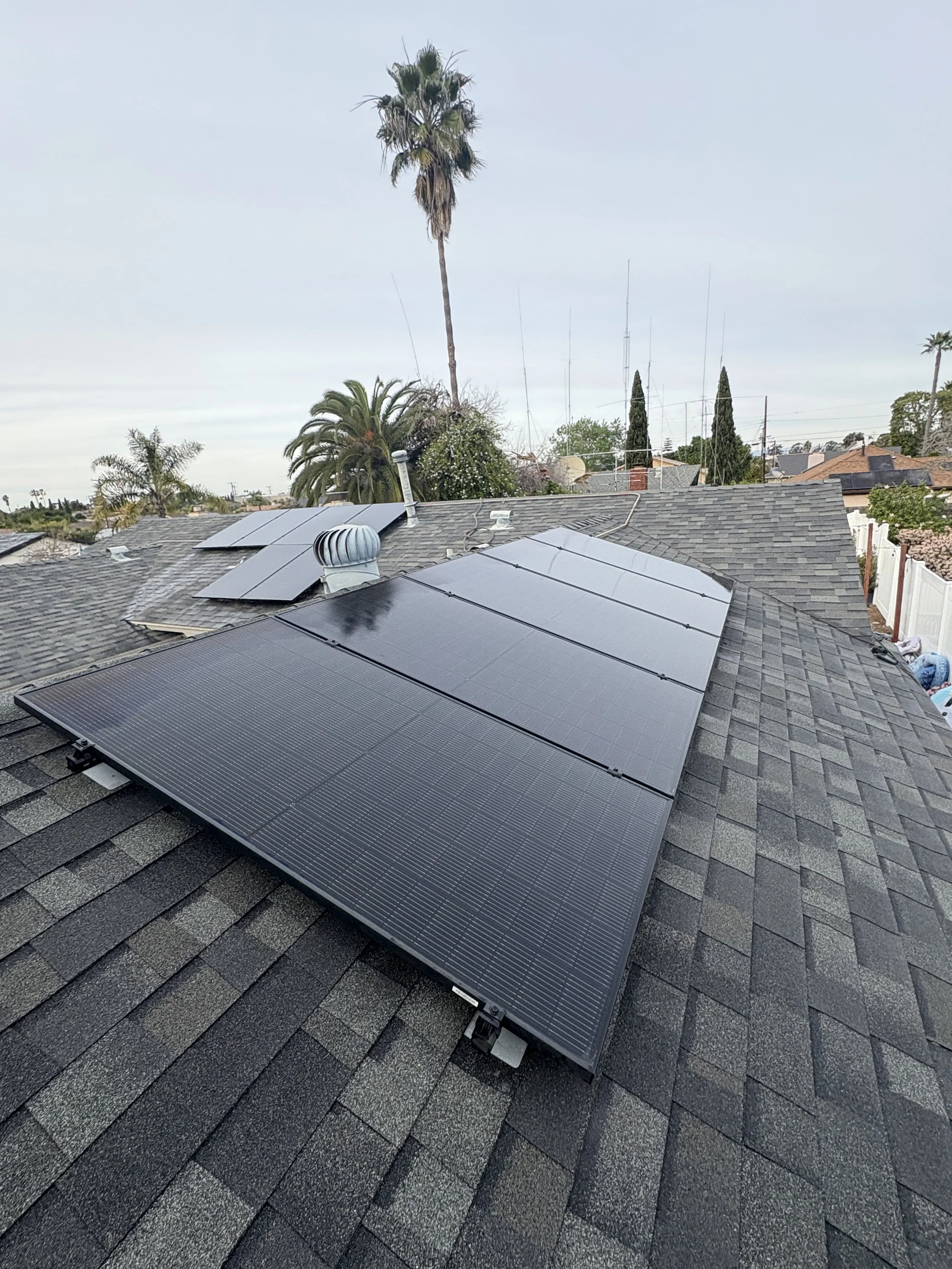 Residential rooftop with black solar panels installed, multiple palm trees in the background, and a cloudy sky.