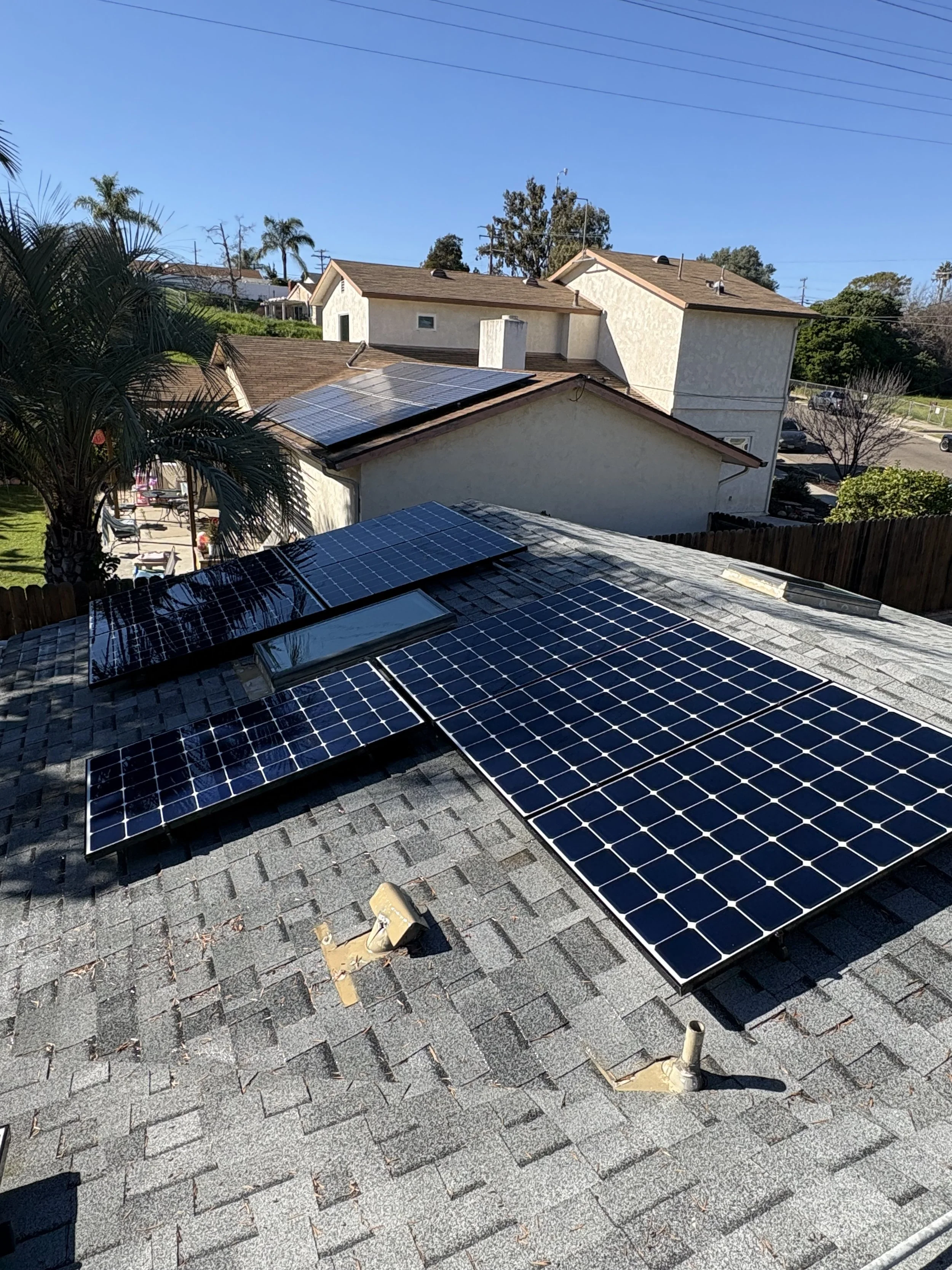Multiple solar panels installed on a house roof, with neighboring houses and blue sky in the background.