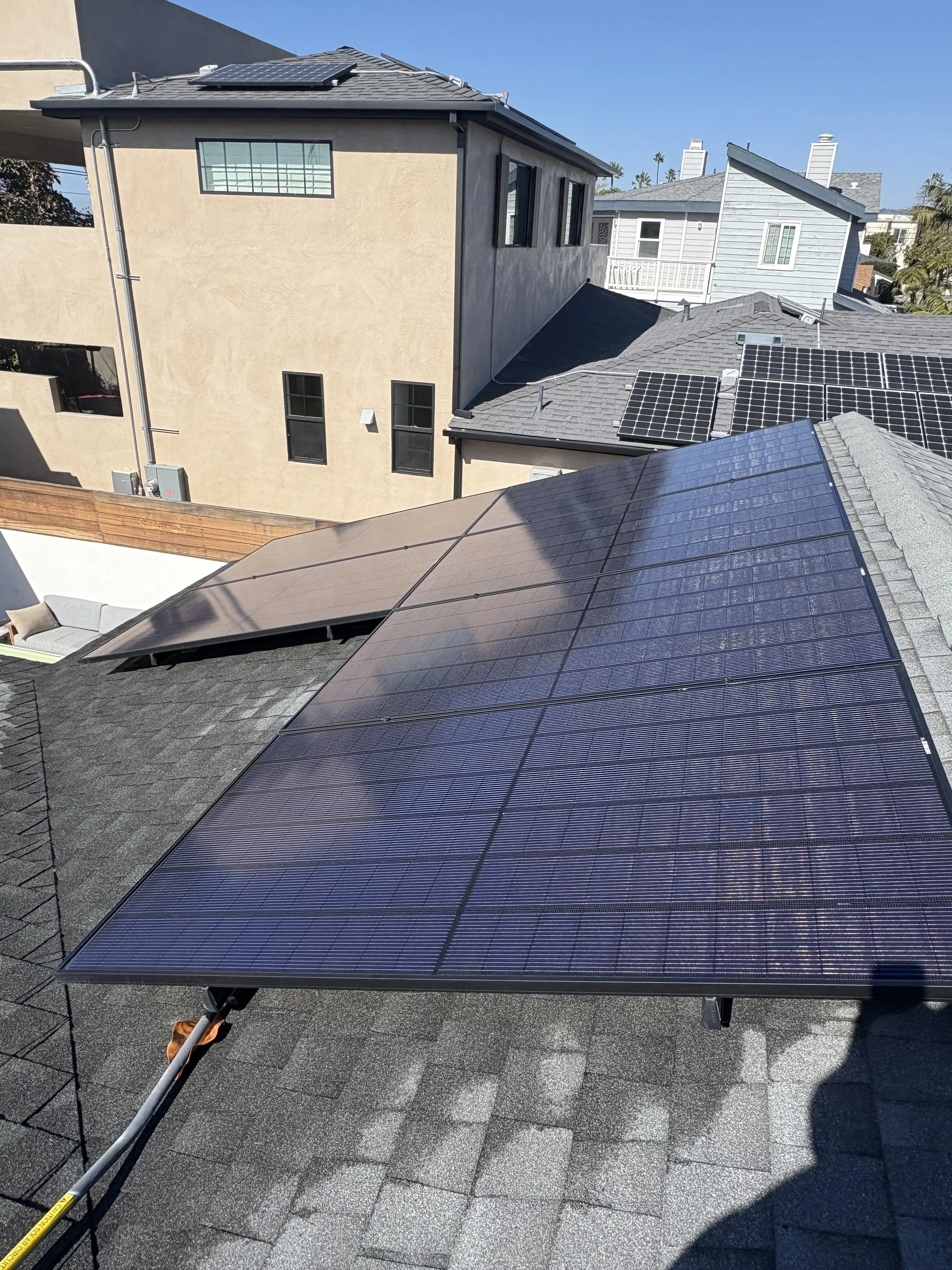 Residential rooftop with newly installed solar panels and neighboring houses with asphalt shingle roofs, some also equipped with solar panels, under a clear blue sky.