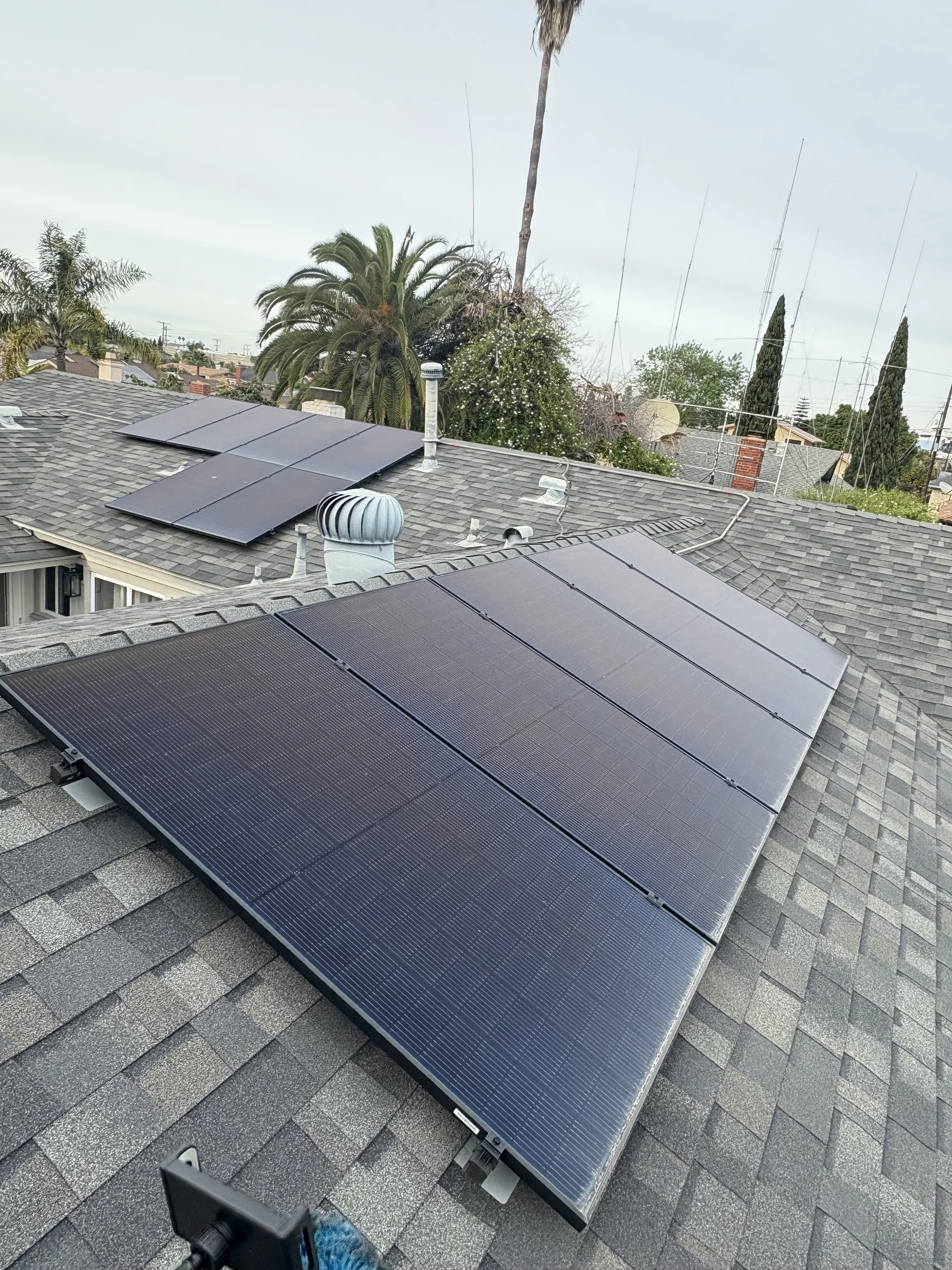 Solar panels installed on residential rooftops in a neighborhood with trees and antennas in the background.