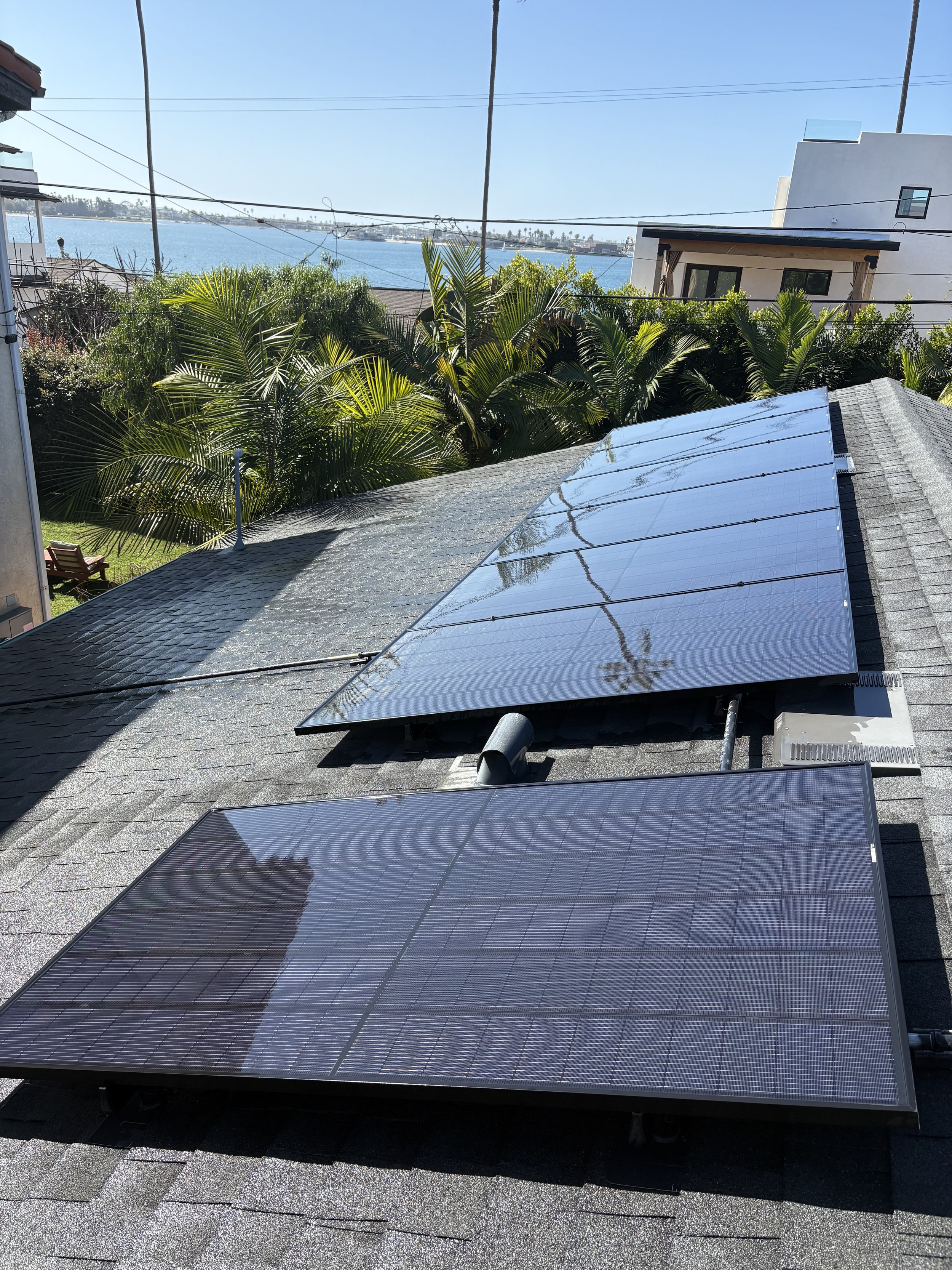 View of a roof with two mounted solar panels, overlooking a backyard with lush green plants and palm trees, and a body of water in the distance under a clear blue sky.