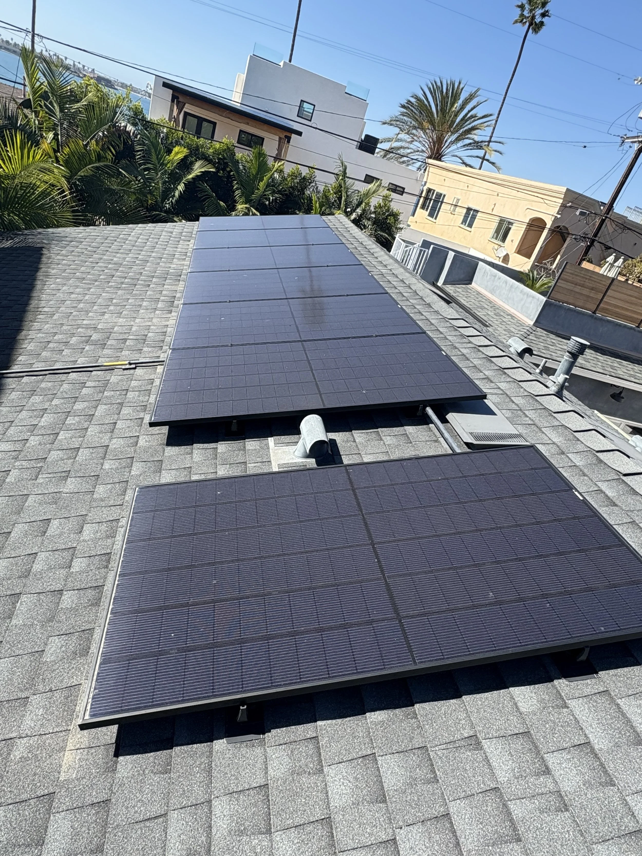 Multiple solar panels installed on a residential rooftop, with modern houses, palm trees, and power lines in the background under clear blue sky.