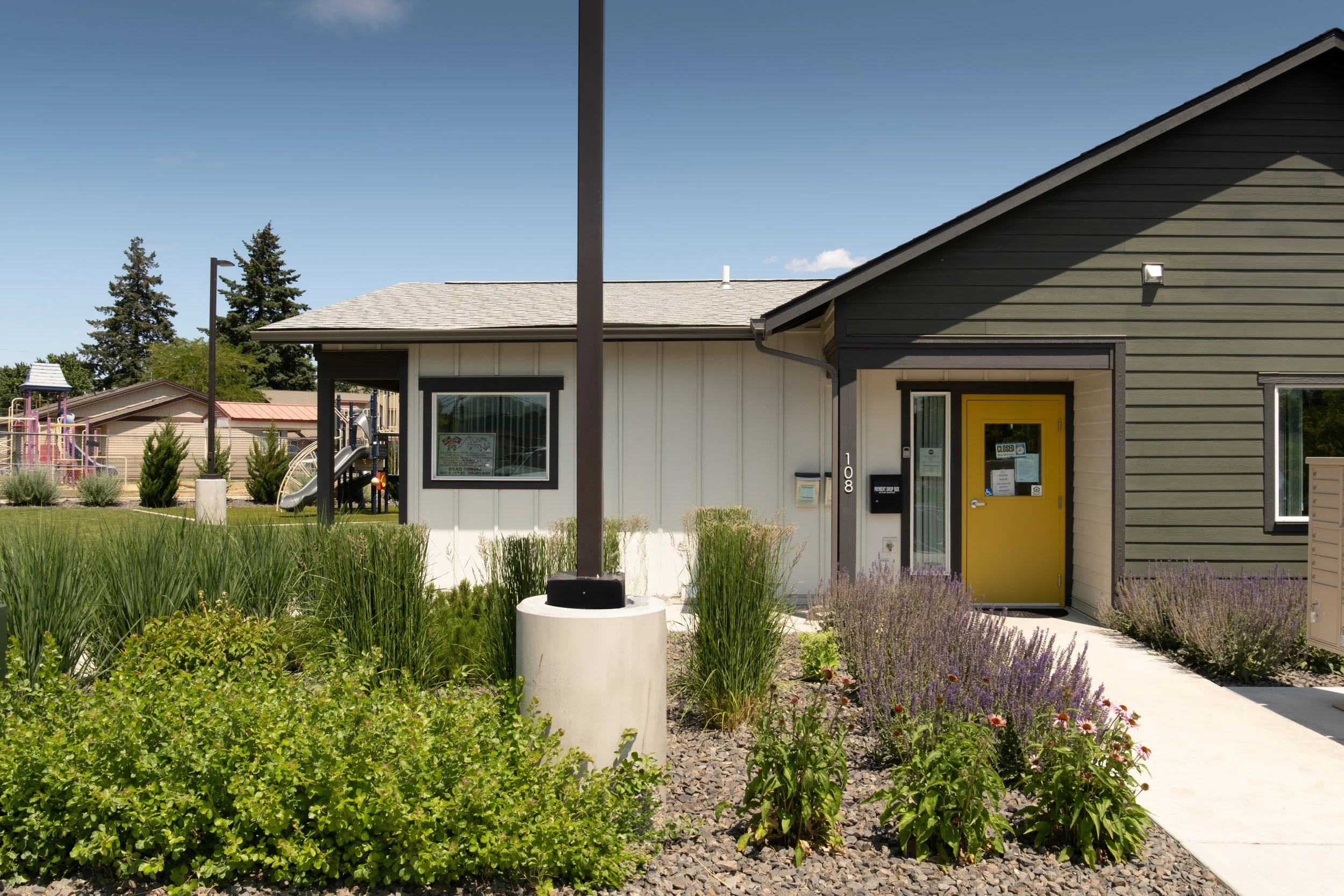 Exterior view of a modern building with a yellow door, gray siding, and a landscaped garden with various plants and shrubs. A sidewalk runs in front of the building, and there are other buildings and playground equipment visible in the background under a clear blue sky.