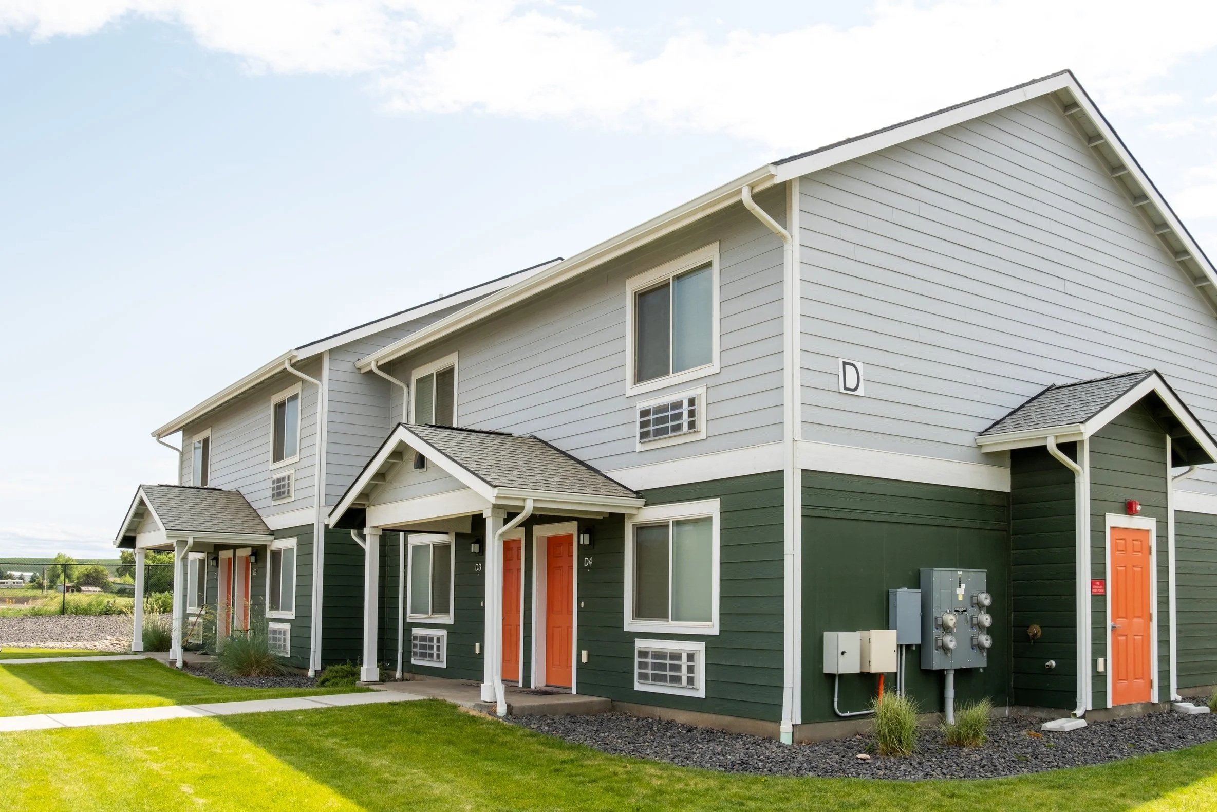 A modern multi-unit apartment building with gray and green siding, orange doors, and small front porches with white columns, set in a well-maintained grassy area under a blue sky.