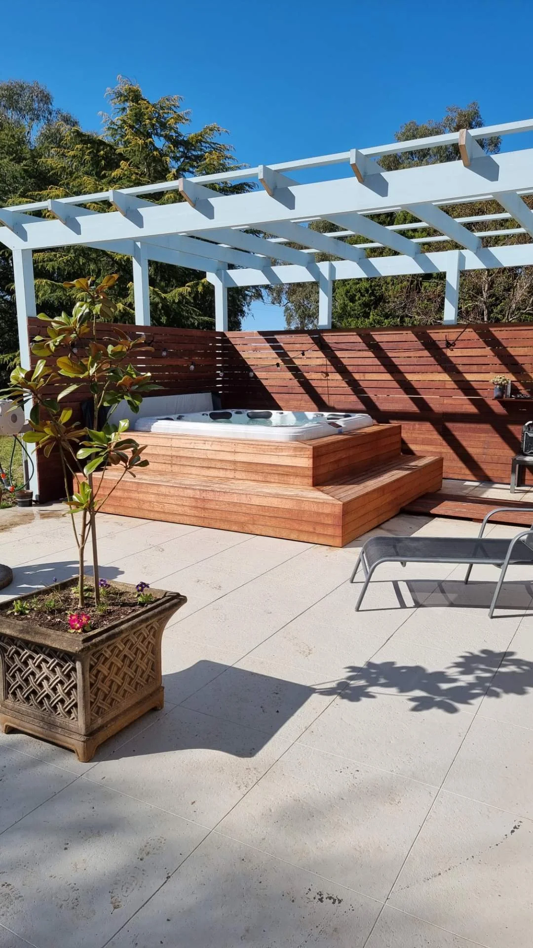 Outdoor patio with a hot tub on a wooden platform, a potted plant, a lounge chair, and a wooden privacy fence with a plant on it, under a partly shaded pergola, bright sunny day with clear blue sky.