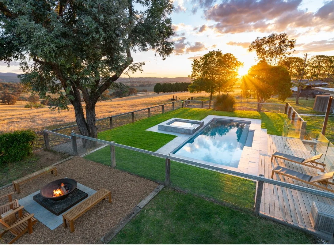 A backyard with a swimming pool, hot tub, and a fire pit area at sunset, surrounded by trees and fields.