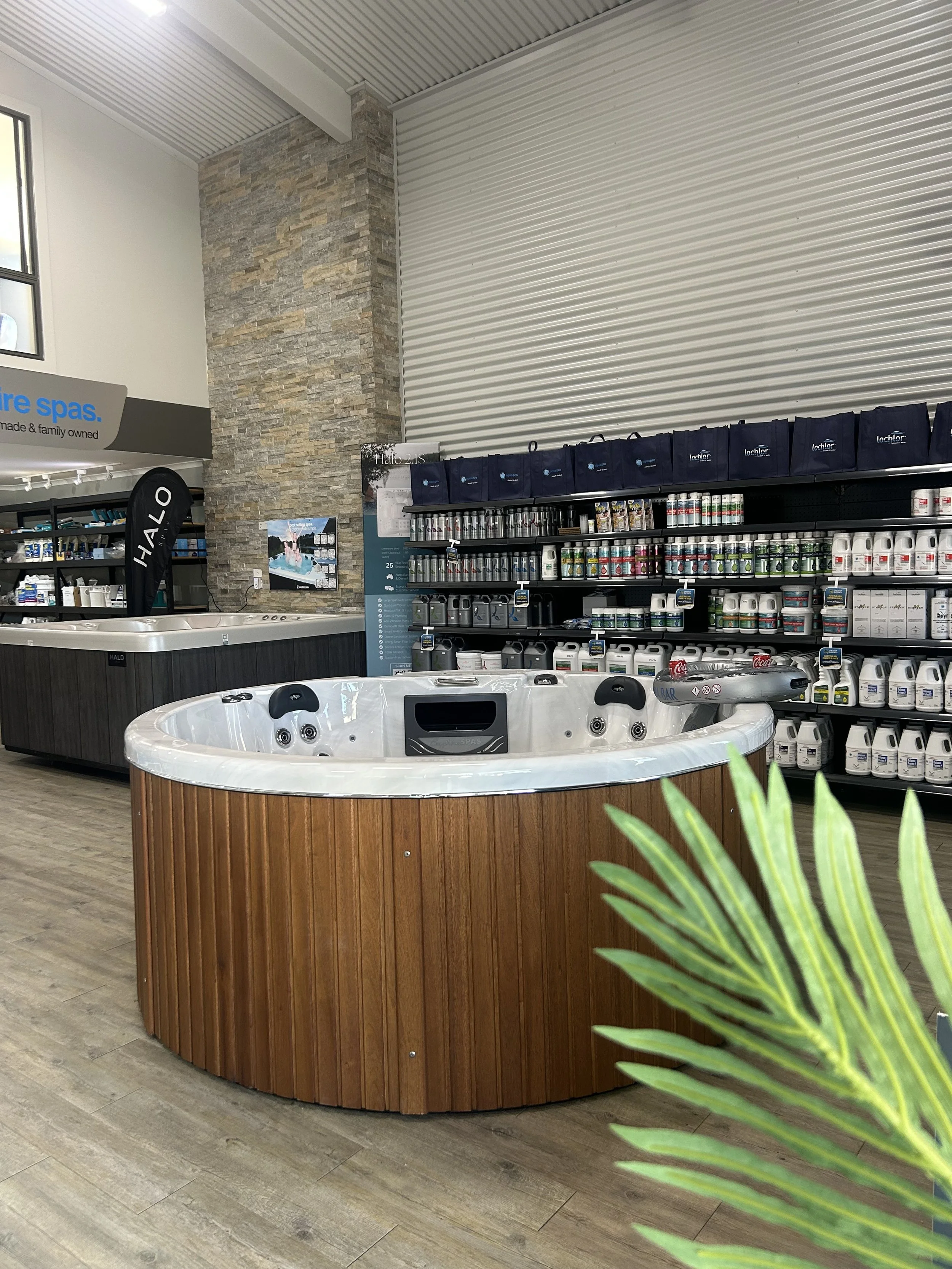 An indoor showroom displaying a large white hot tub with a wooden exterior panel, surrounded by shelves stocked with pool chemicals and accessories, with a stone pillar and a wall with corrugated metal paneling in the background.