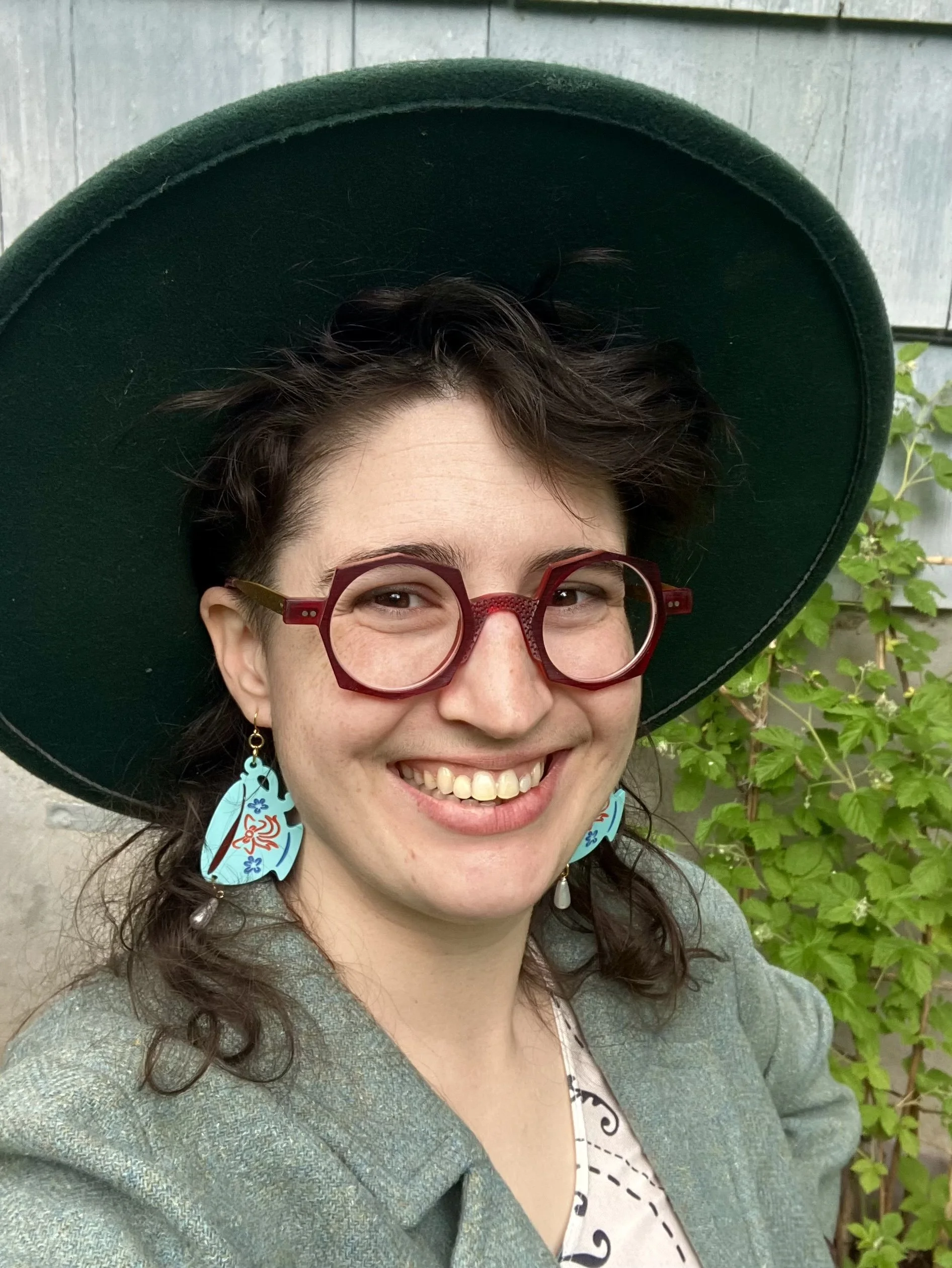 A smiling person with short dark hair, red glasses, teal tea cup earrings, wearing a wide-brimmed green hat, and a gray blazer, outdoors with green foliage in the background.