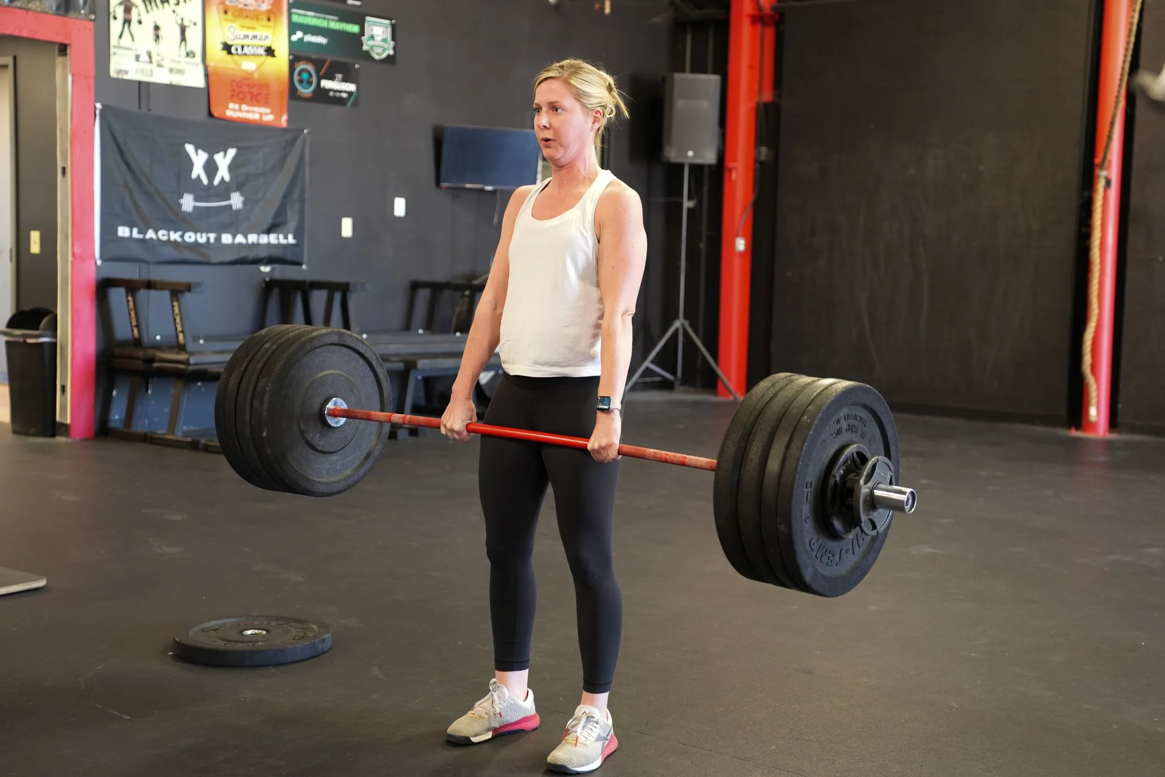 A woman assists another woman during a physical therapy or fitness session in a gym, with exercise equipment visible in the background.