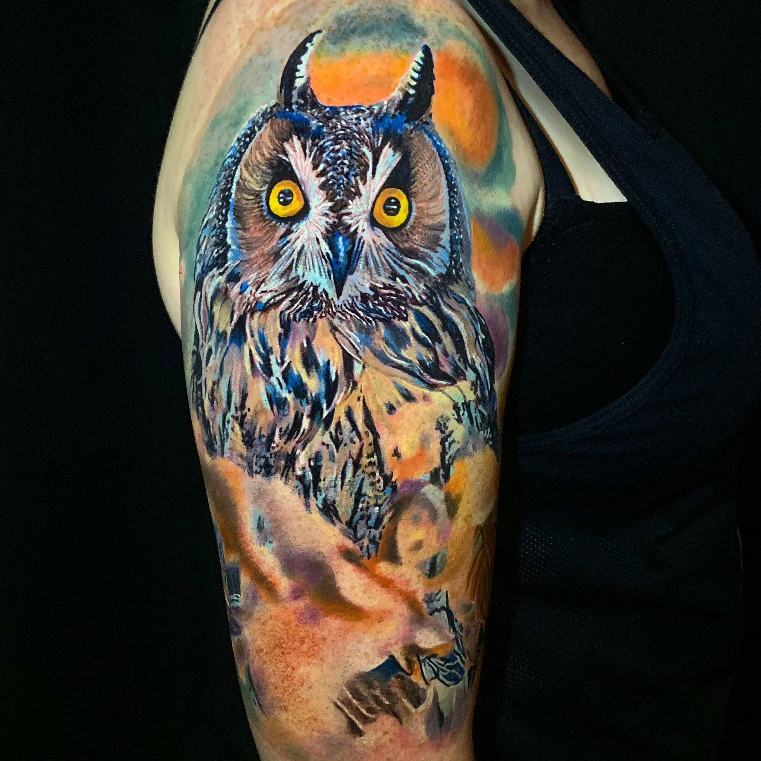 Colorful tattoo of an owl with yellow eyes on a person's arm, with a blurred background.