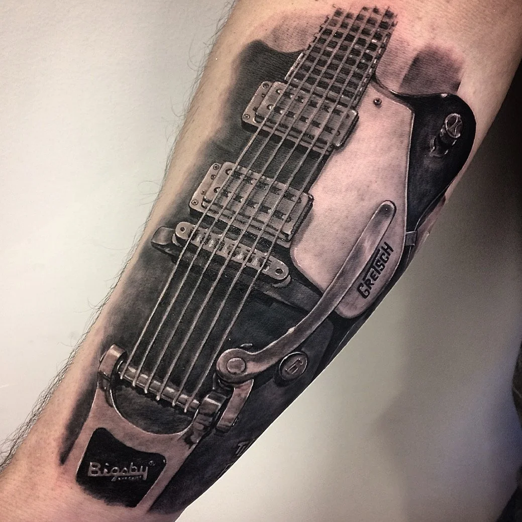 Tattoo of a realistic electric guitar on a person's arm, featuring details like pickups, strings, a tremolo arm with the word 'GRESH,' and the brand name 'Bigsby' on the tailpiece.