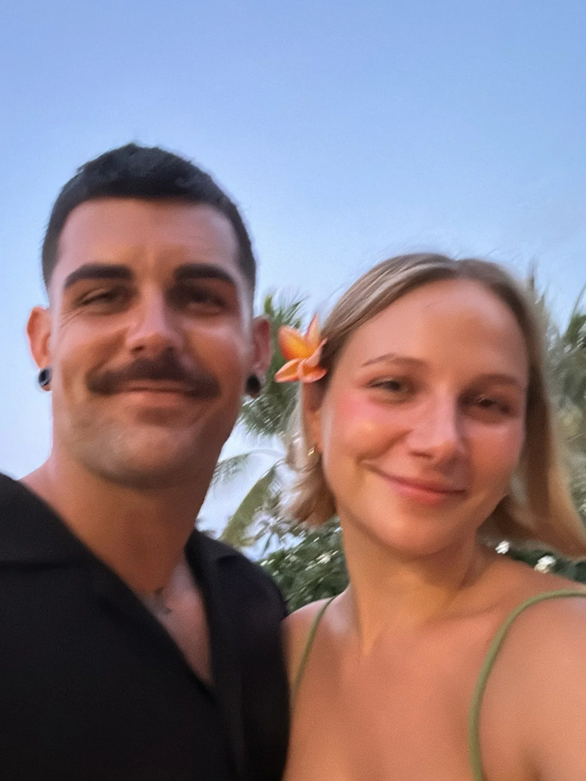 A young man with dark hair and a moustache and earring, and a young woman with blond hair and an orange flower in her hair, smiling together outdoors with palm trees in the background during sunset.