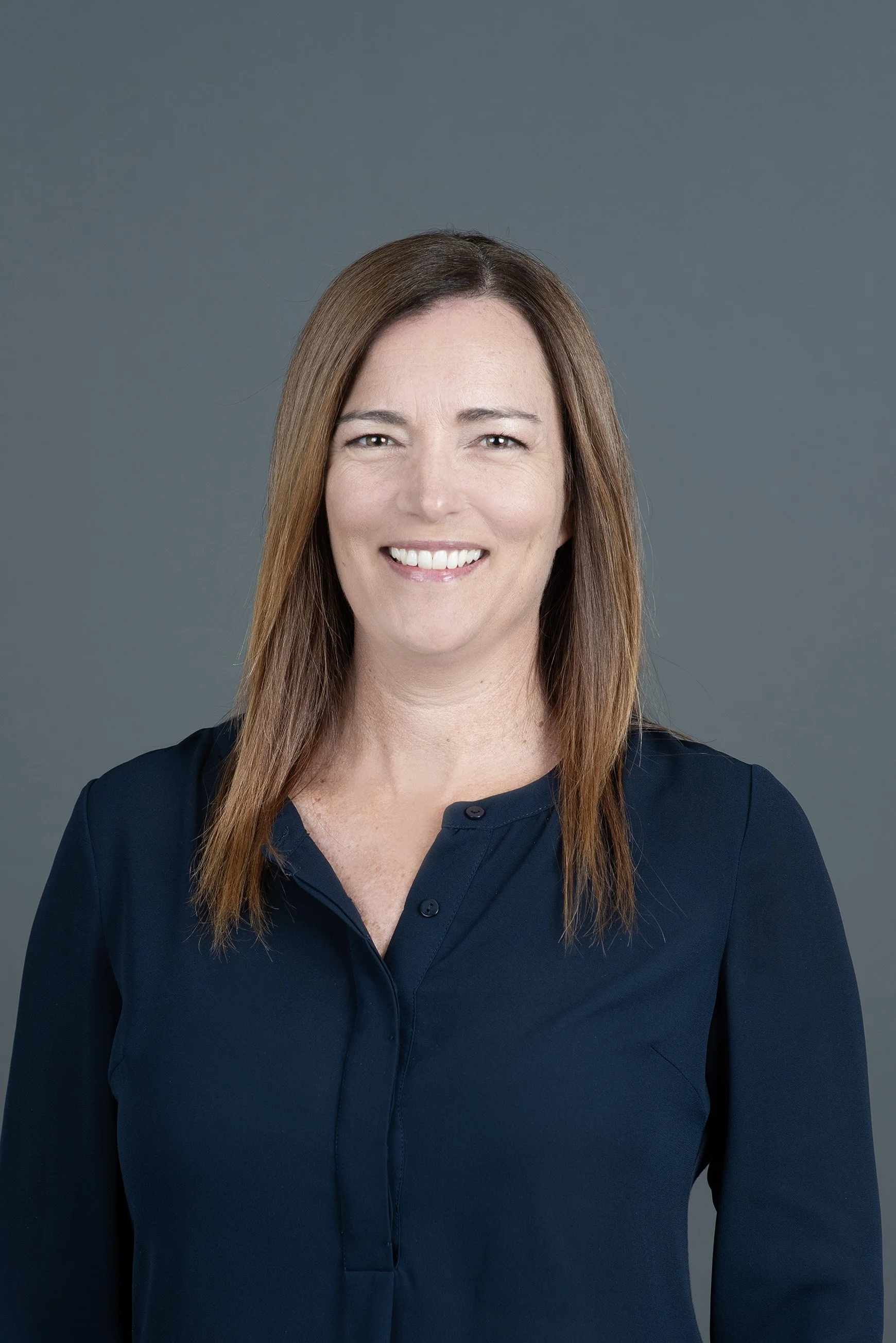 Smiling woman with long reddish-brown hair wearing a black blazer, facing the camera against a plain background.