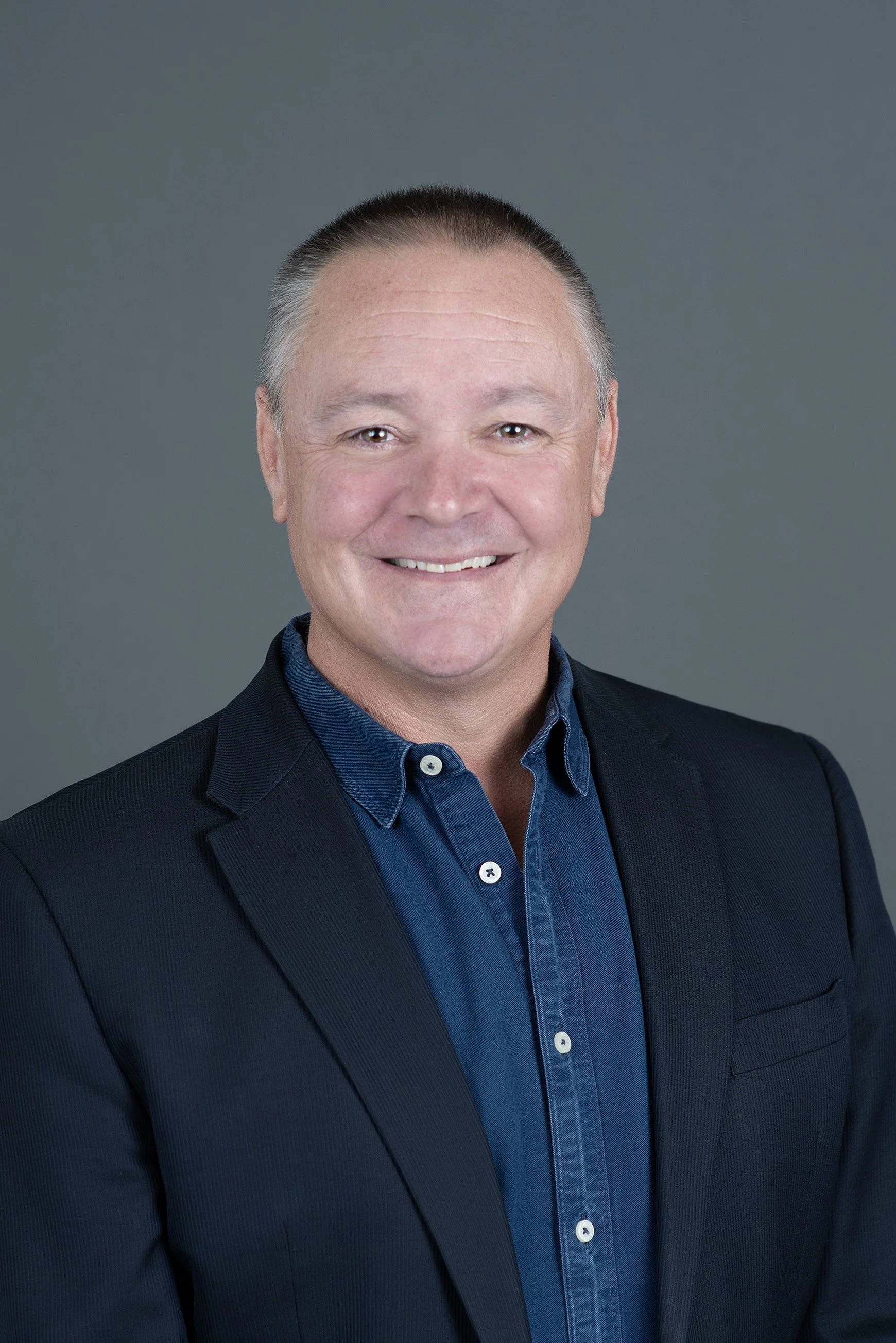 A smiling middle-aged man with short gray hair, wearing a dark blazer over a light blue collared shirt, against a neutral background.