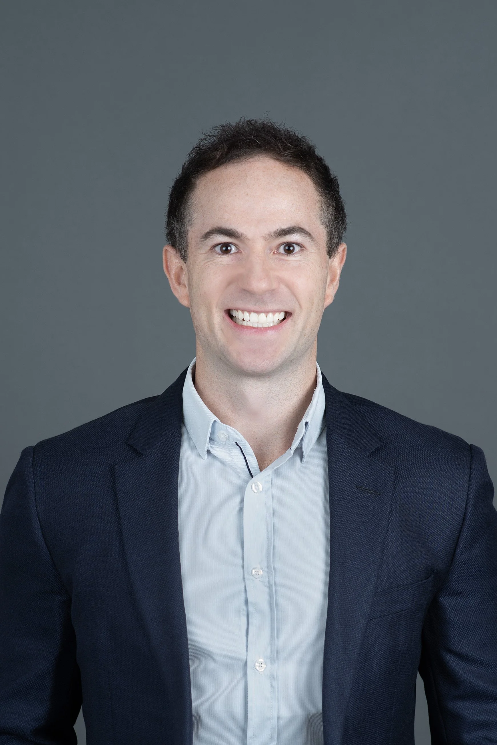 Portrait of a man wearing a light pink button-down shirt smiling at the camera against a plain white background.