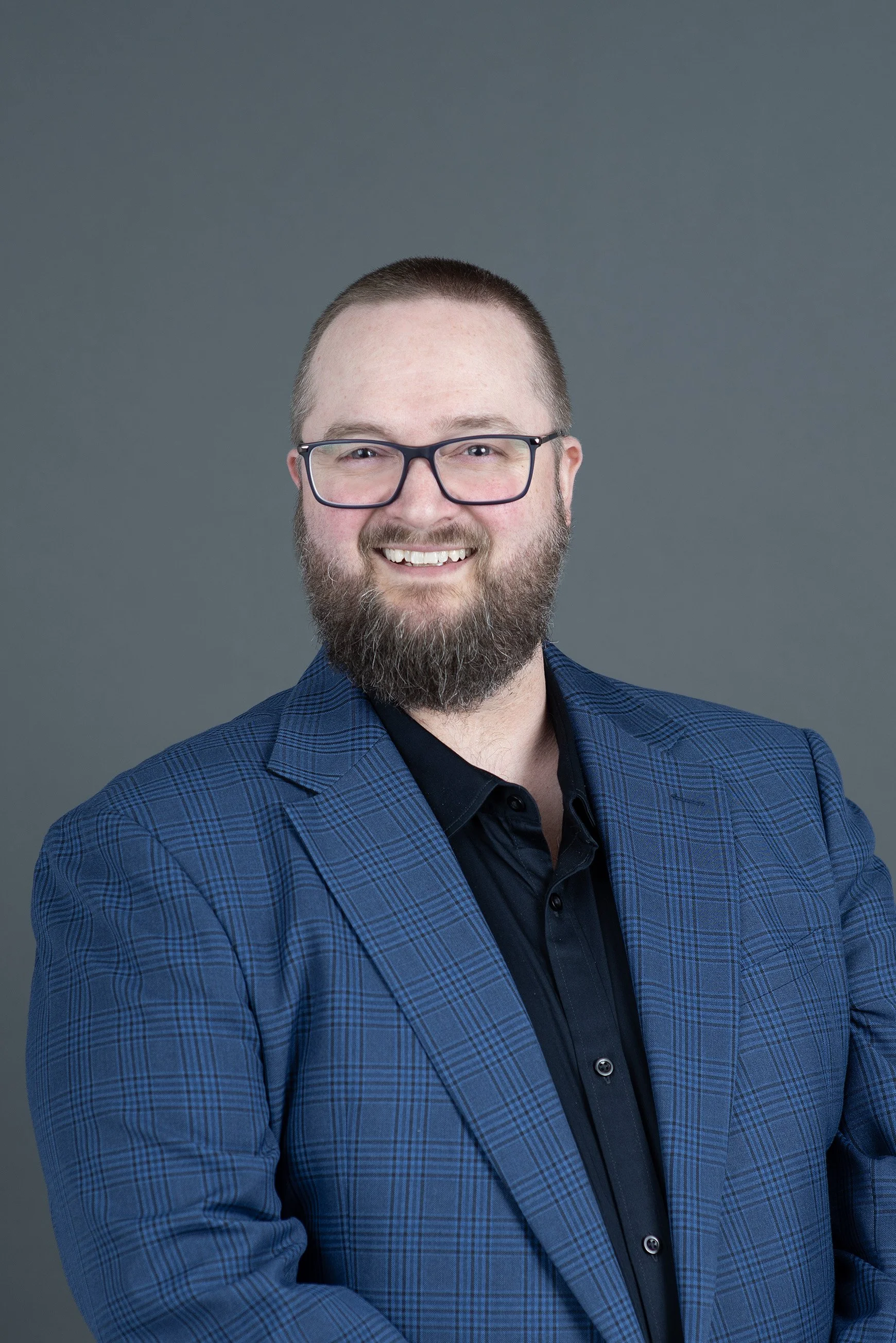 A man with glasses, a beard, and short hair, wearing a blue collared shirt, smiling at the camera against a gray background.