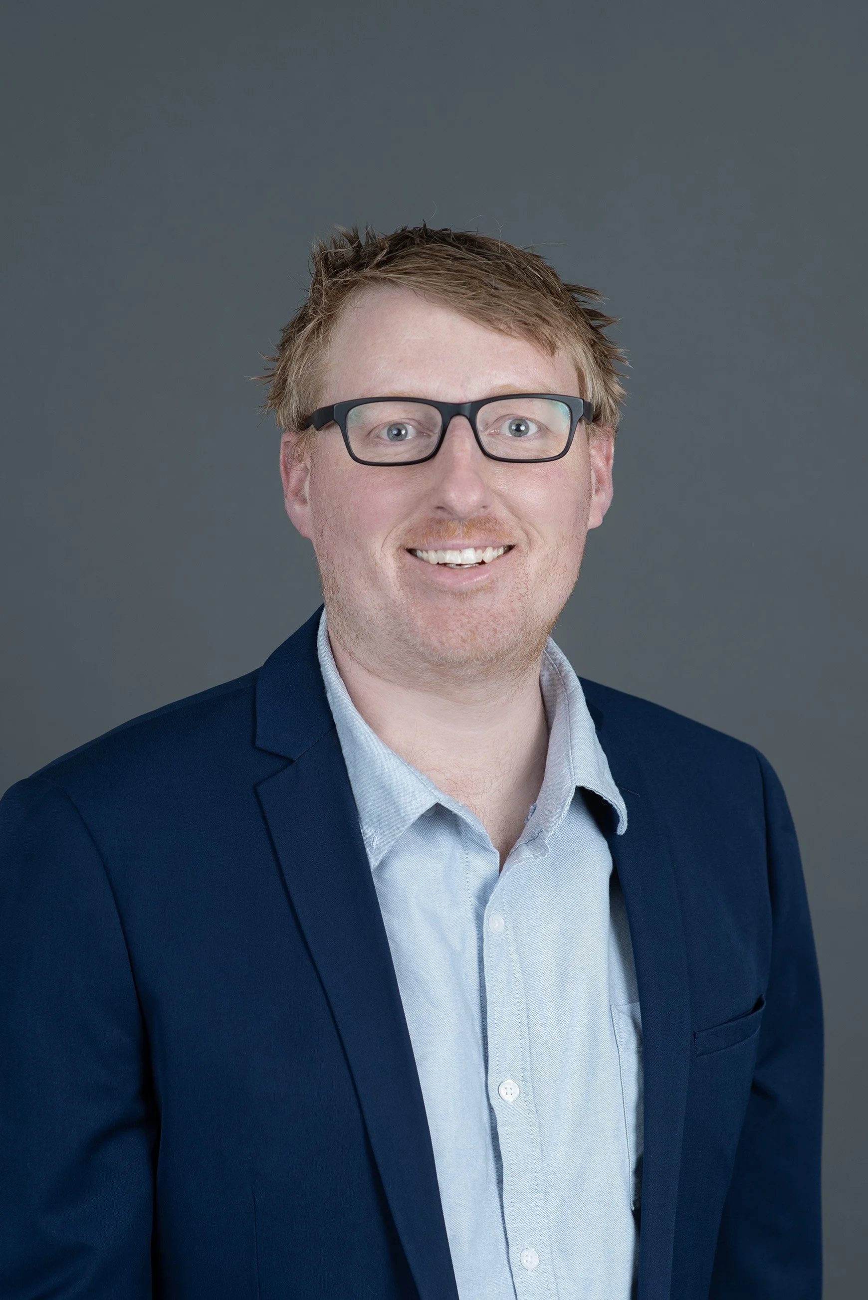 A man with light skin, short messy blonde hair, glasses, and a partial beard smiling at the camera, wearing a blue collared shirt against a plain gray background.