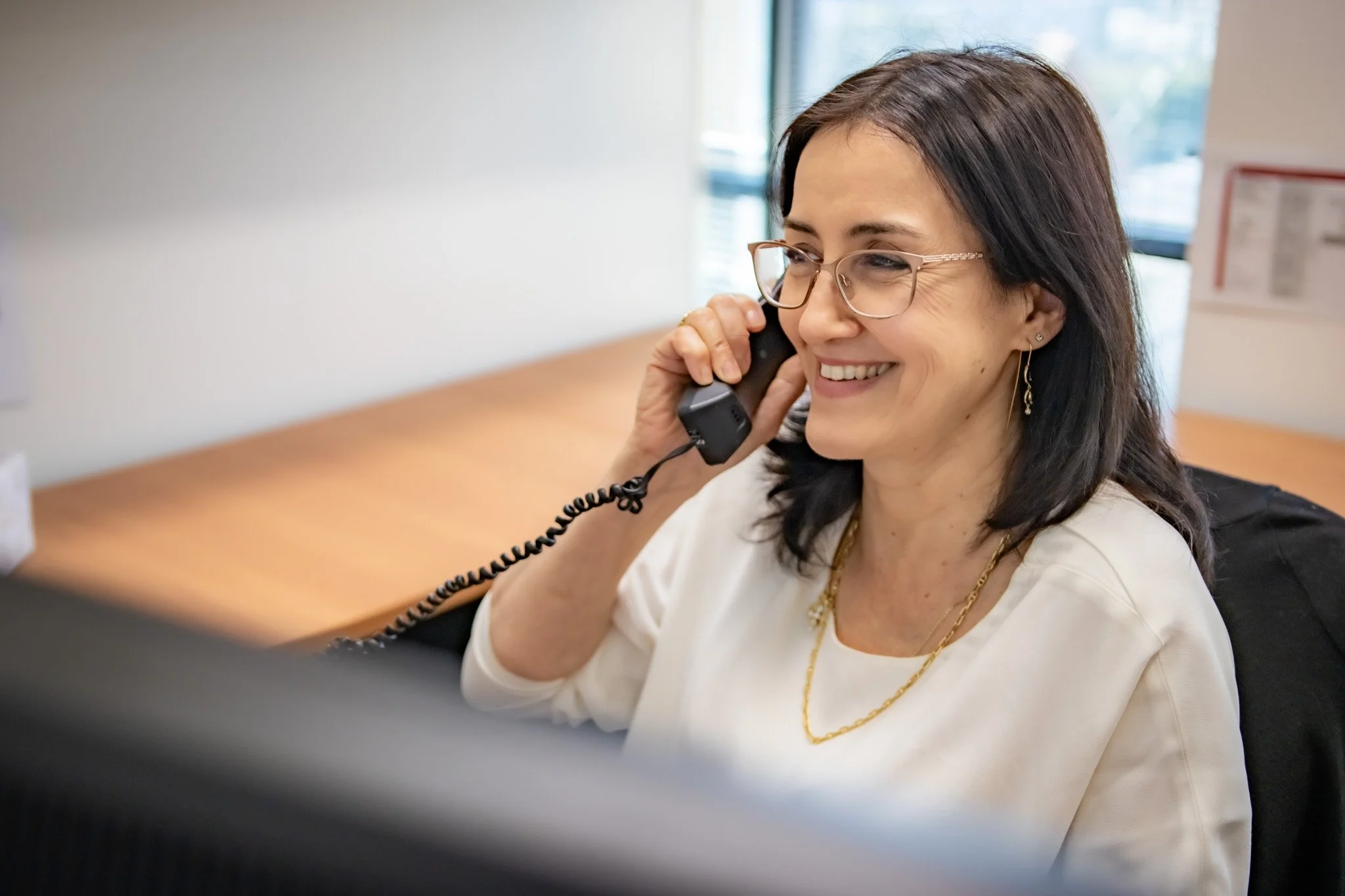 A woman with dark hair and glasses smiling while talking on a landline phone in an office setting.