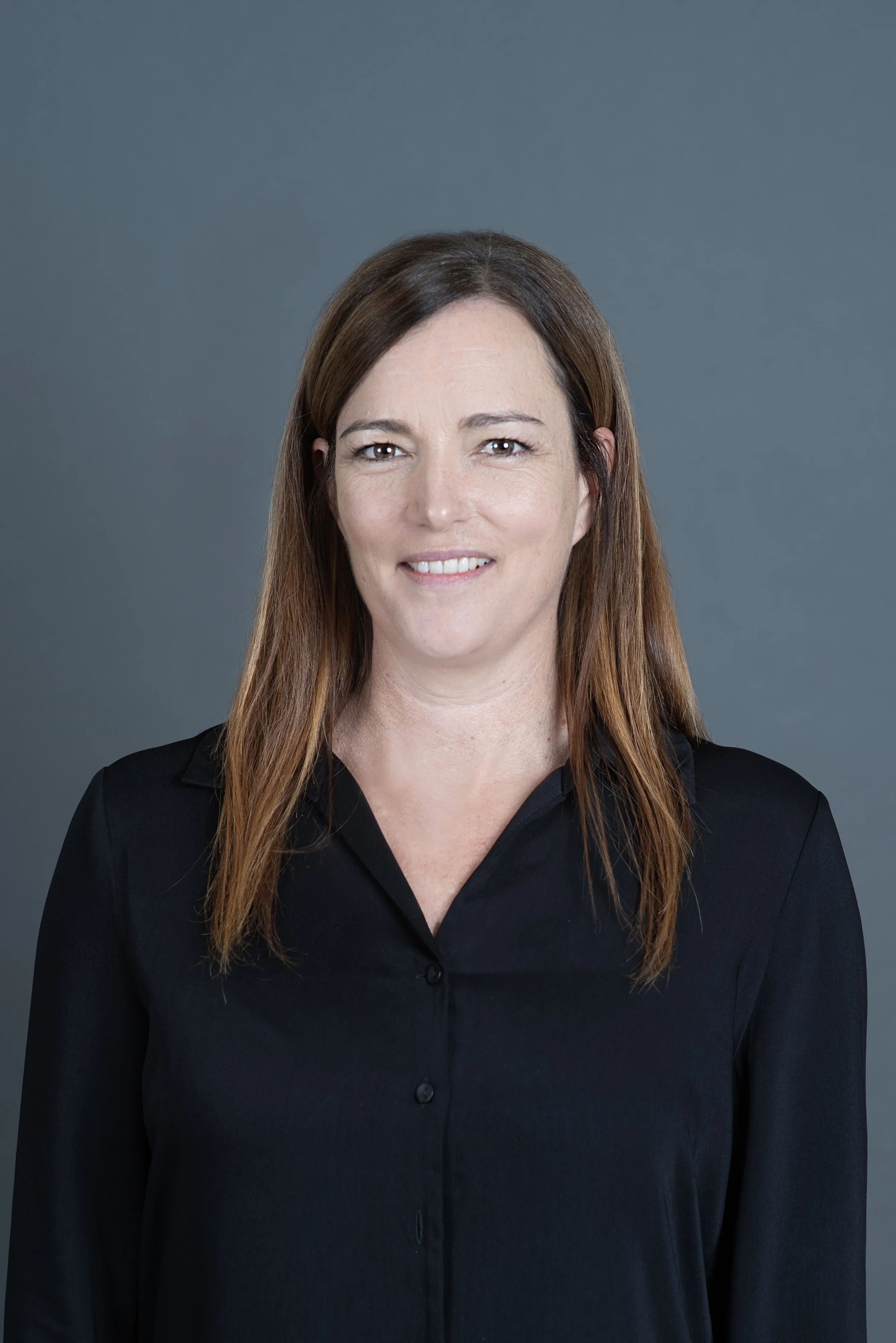 Smiling woman with long reddish-brown hair wearing a black blazer, facing the camera against a plain background.