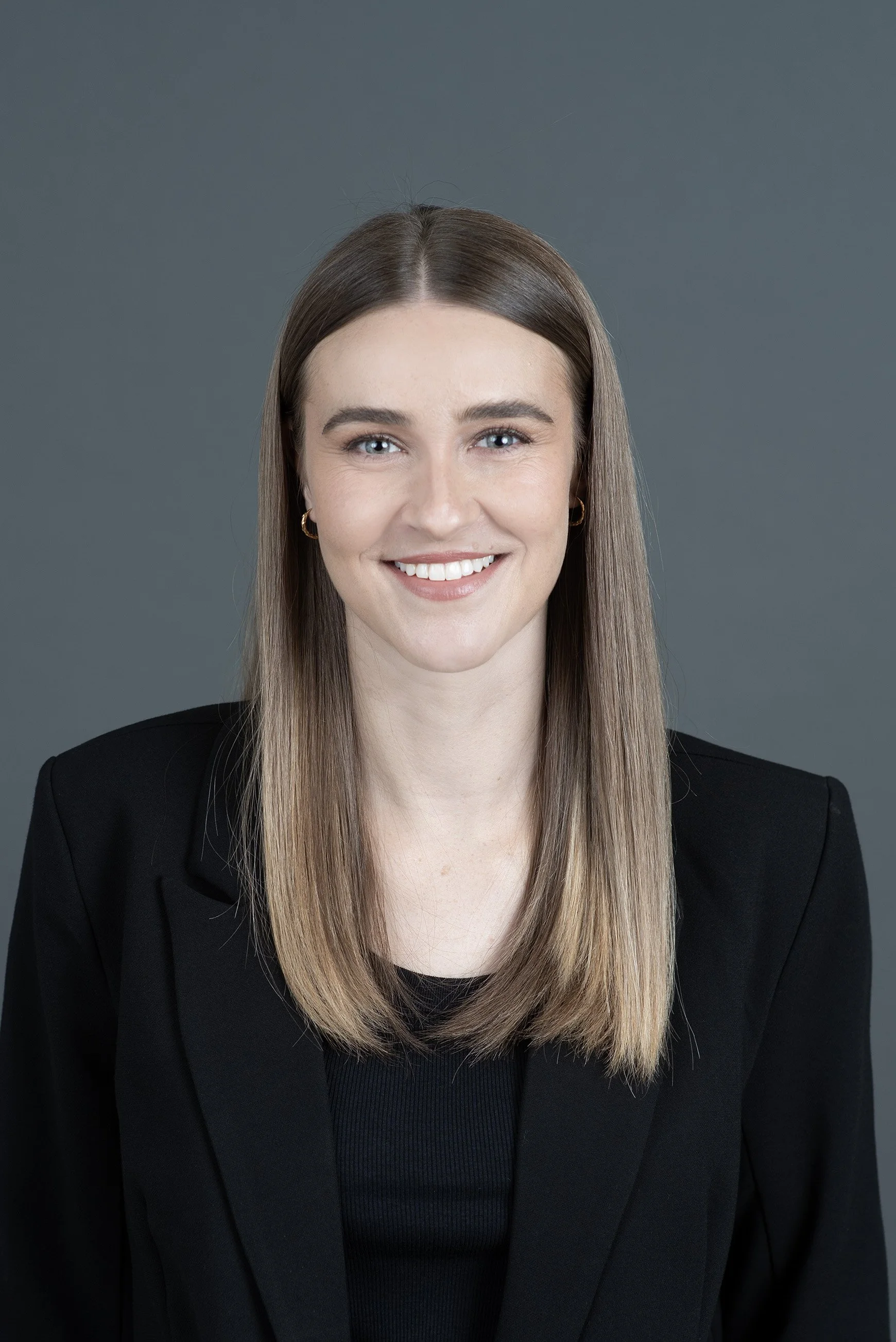 A woman with long, straight, light brown hair smiling at the camera against a plain grey background.