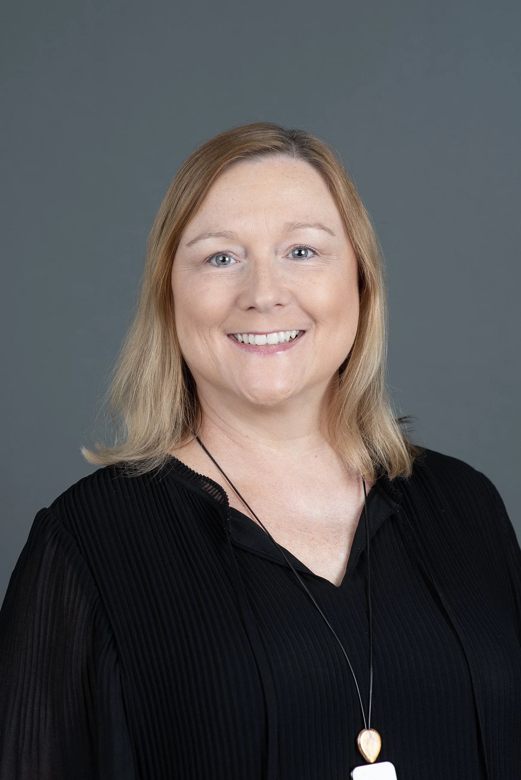 A woman with blonde hair smiling at the camera, wearing a pink top, against a plain light background.