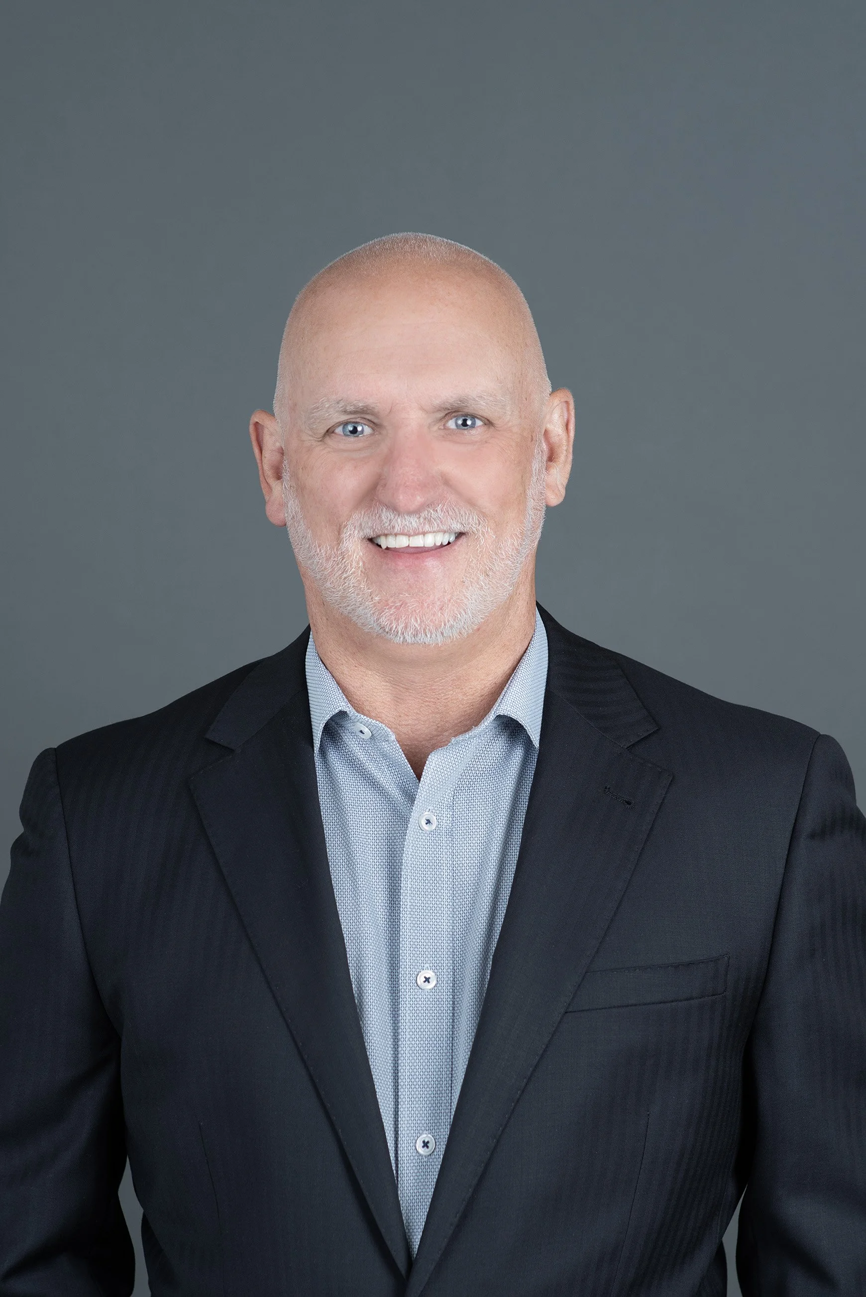 Headshot of a bald man with blue eyes, smiling, wearing a dark blue button-up shirt against a light gray background.