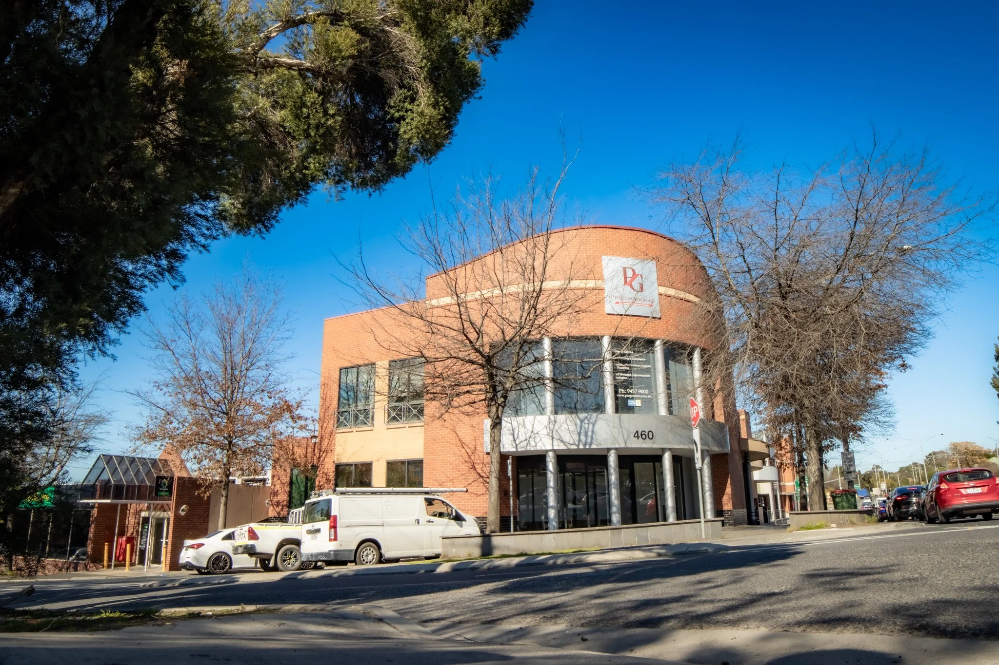 A two-story brick commercial building with large glass windows and metal pillars, situated on a street with parked cars and leafless trees, under a clear blue sky.