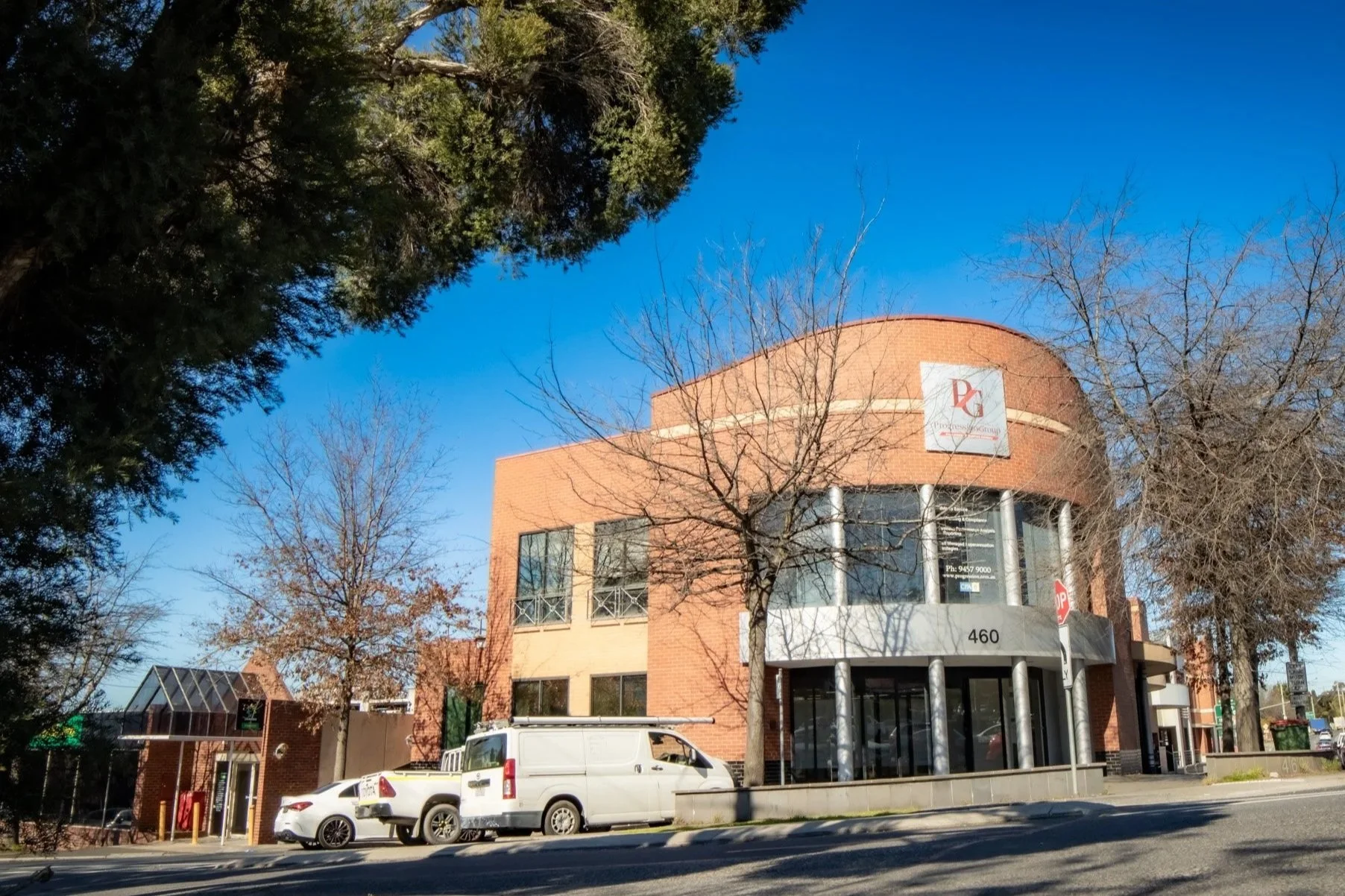 A modern brick building with large glass windows and a curved facade. The building has a sign on it displaying a logo with the letters 'PG'. Several parked cars, including a white van and a white SUV, are in front of the building. The street is lined with leafless trees, indicating a fall or winter season. The sky is clear and blue.