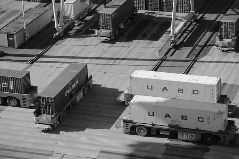 Cargo containers and train cars on a rail yard or port, with some containers on flatbed railcars and others on train cars, in black and white.