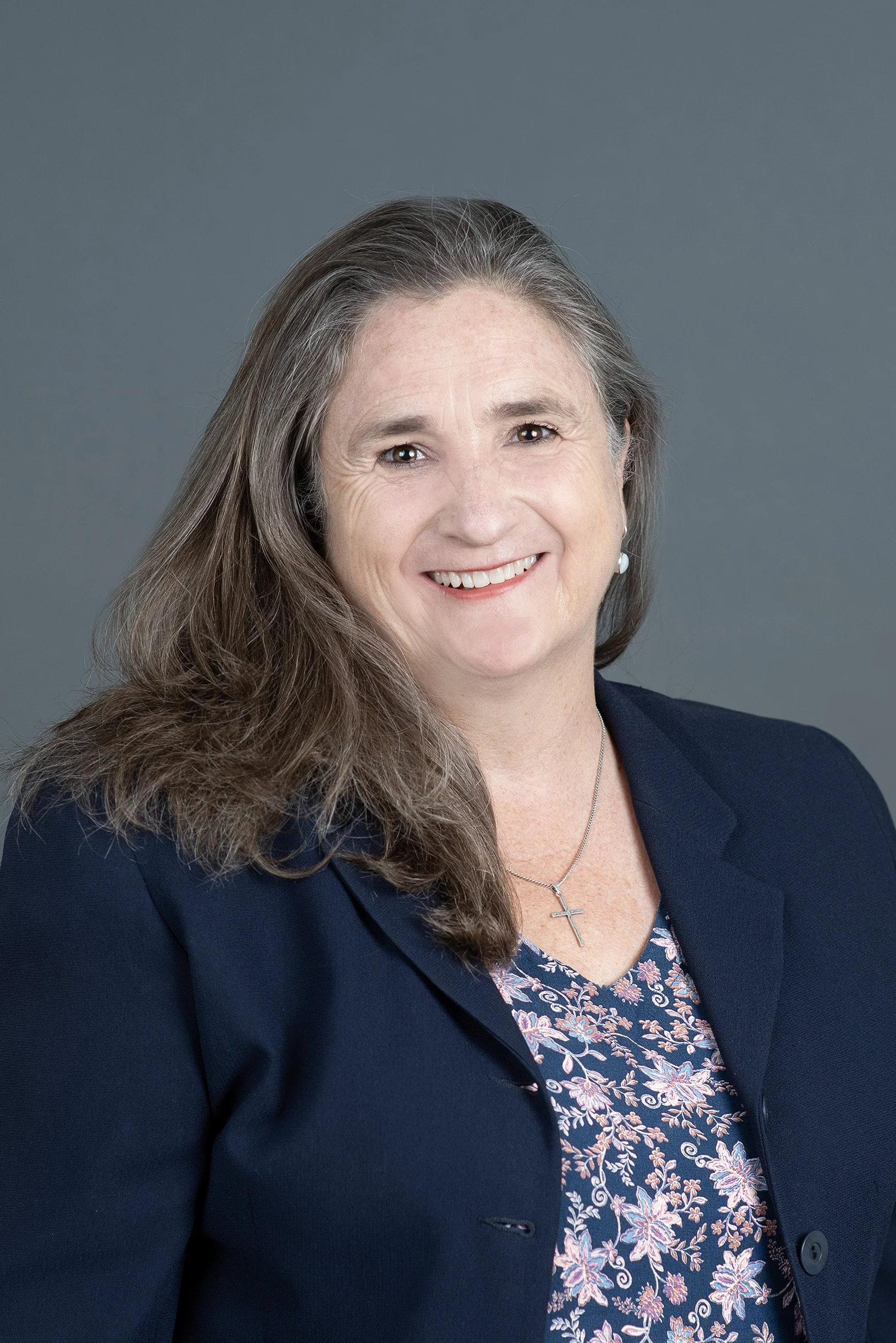 A smiling woman with shoulder-length gray hair, wearing a black and white patterned top and pearl earrings, against a plain gray background.