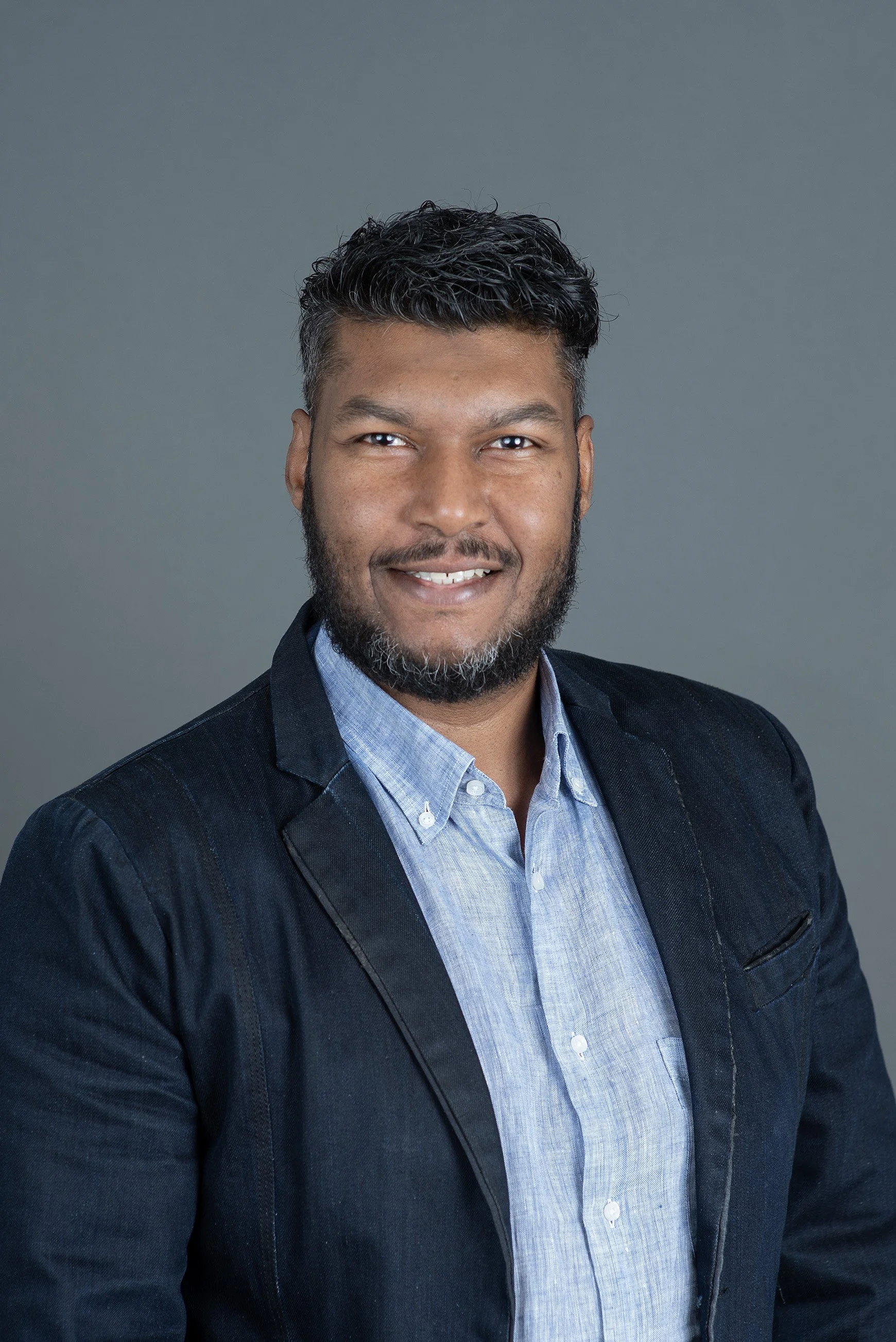 Portrait of a man with curly hair and a beard, wearing a dark blazer and white shirt, against a plain light background.