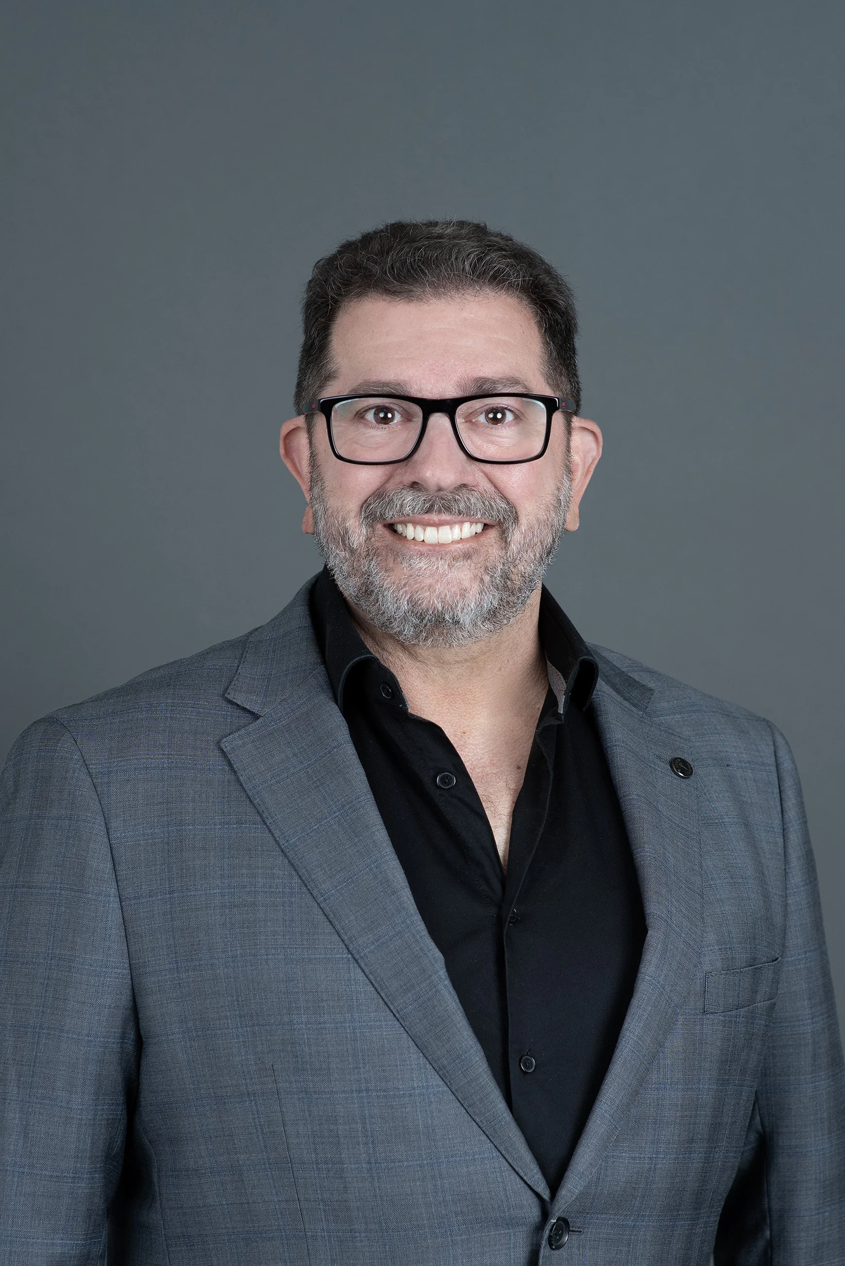 A middle-aged man with short, dark curly hair and a gray beard, wearing a gray suit jacket and black shirt, smiling confidently against a plain white background.