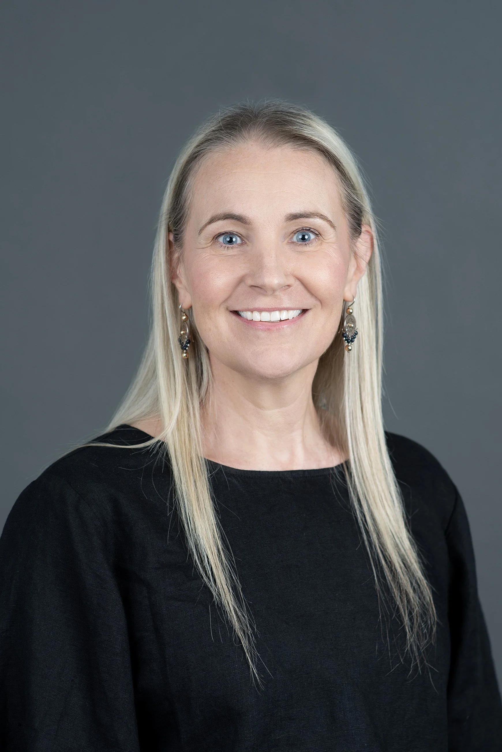 A middle-aged woman with long blonde hair, smiling, wearing earrings and a dark top, posing against a plain white background.