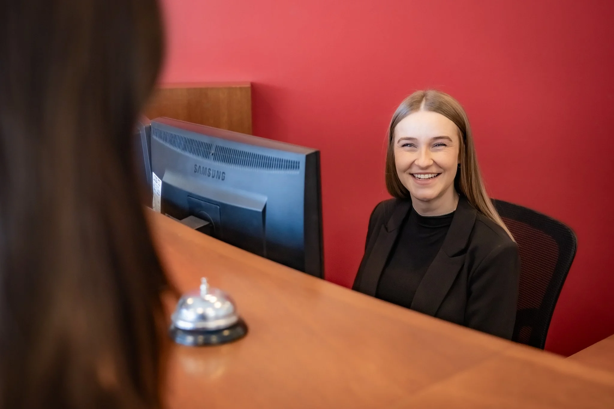 A young woman with long light brown hair smiling at a receptionist behind a desk. The woman is blurred in the foreground, and the background has a red wall.