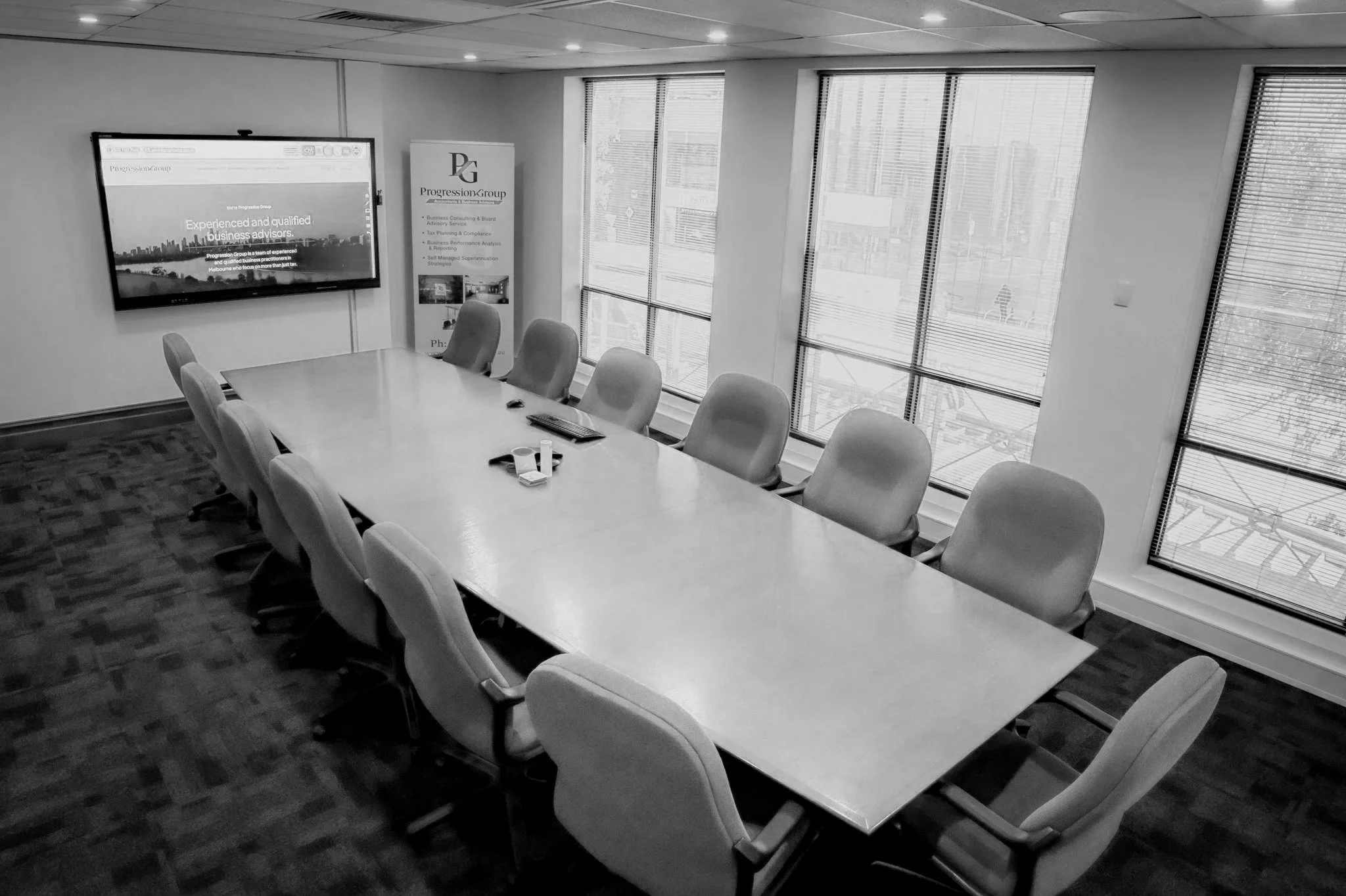 Empty conference room with a long table, chairs, a wall-mounted monitor displaying a website, and large windows with blinds.