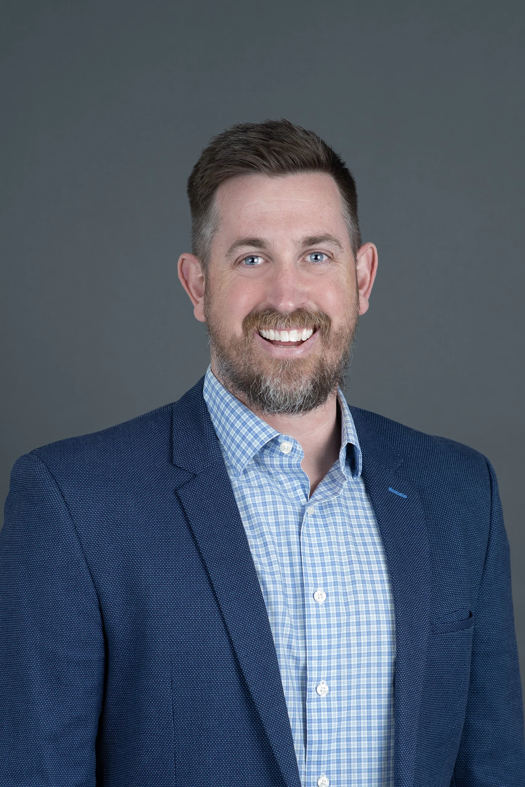 A smiling man with short brown hair, a beard, and blue eyes, wearing a blue checkered shirt against a plain white background.