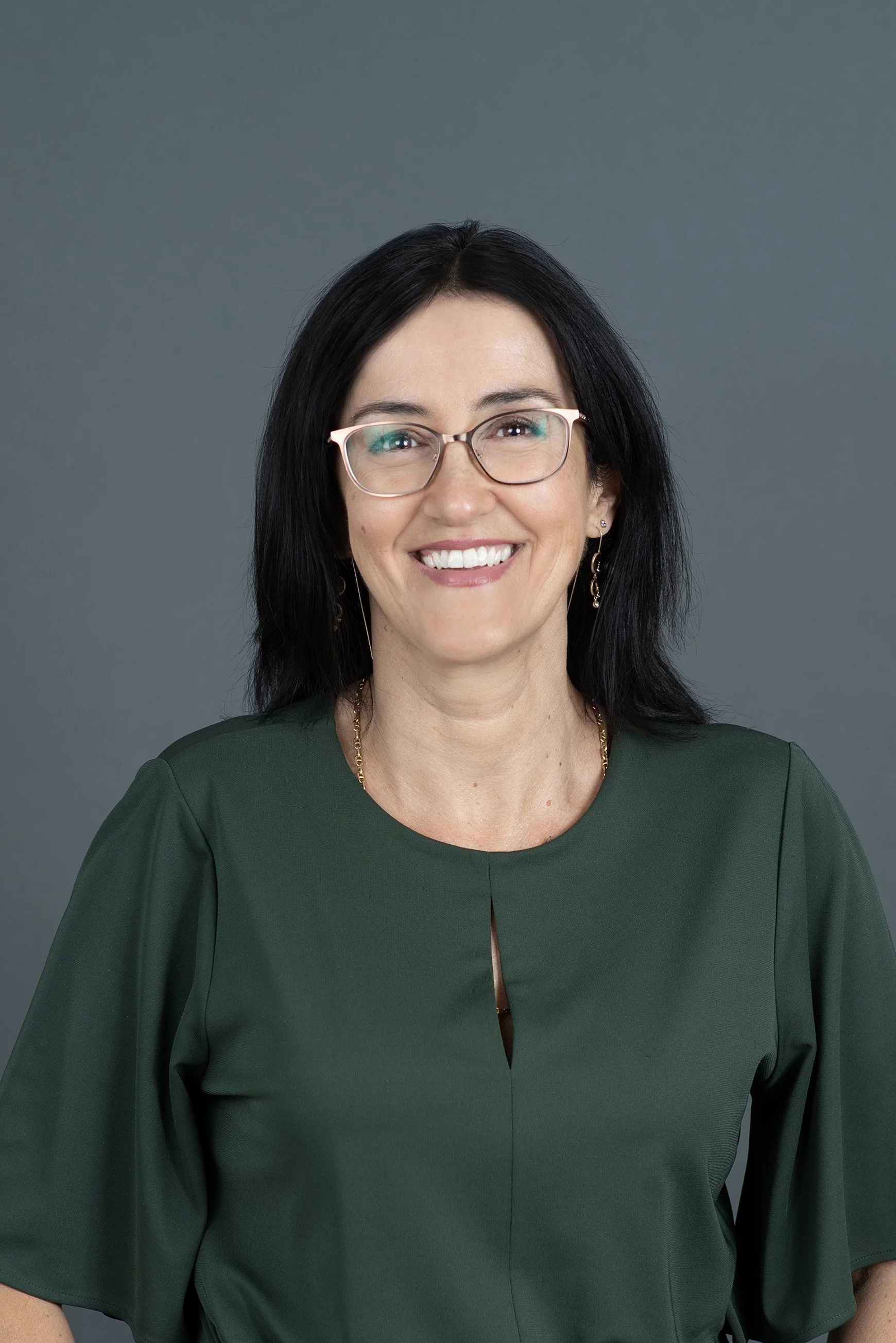 A woman with shoulder-length dark hair, glasses, and gold jewelry smiling at the camera against a plain light background.