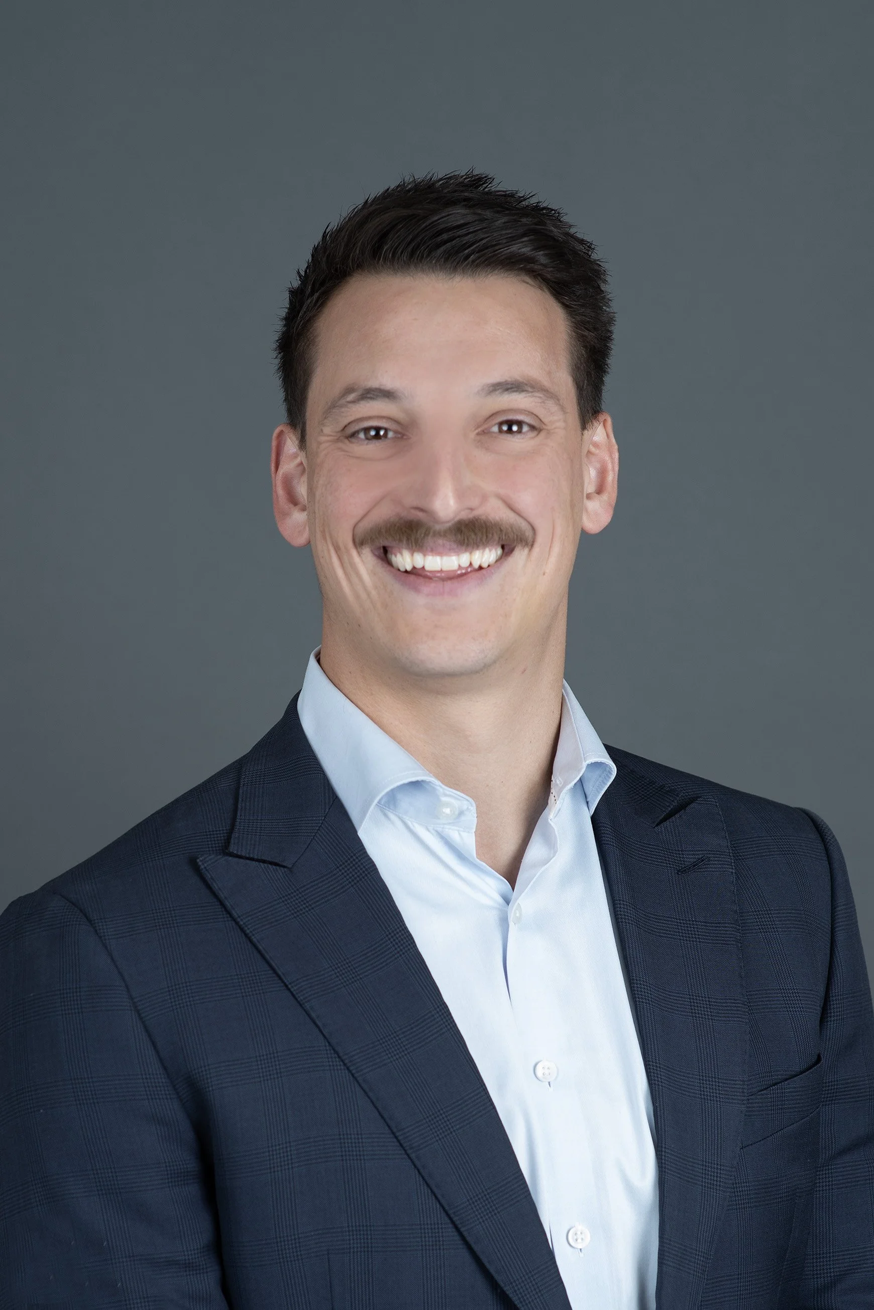 A man with brown hair, a mustache, and light skin smiling, wearing a light blue button-up shirt, against a plain light background.