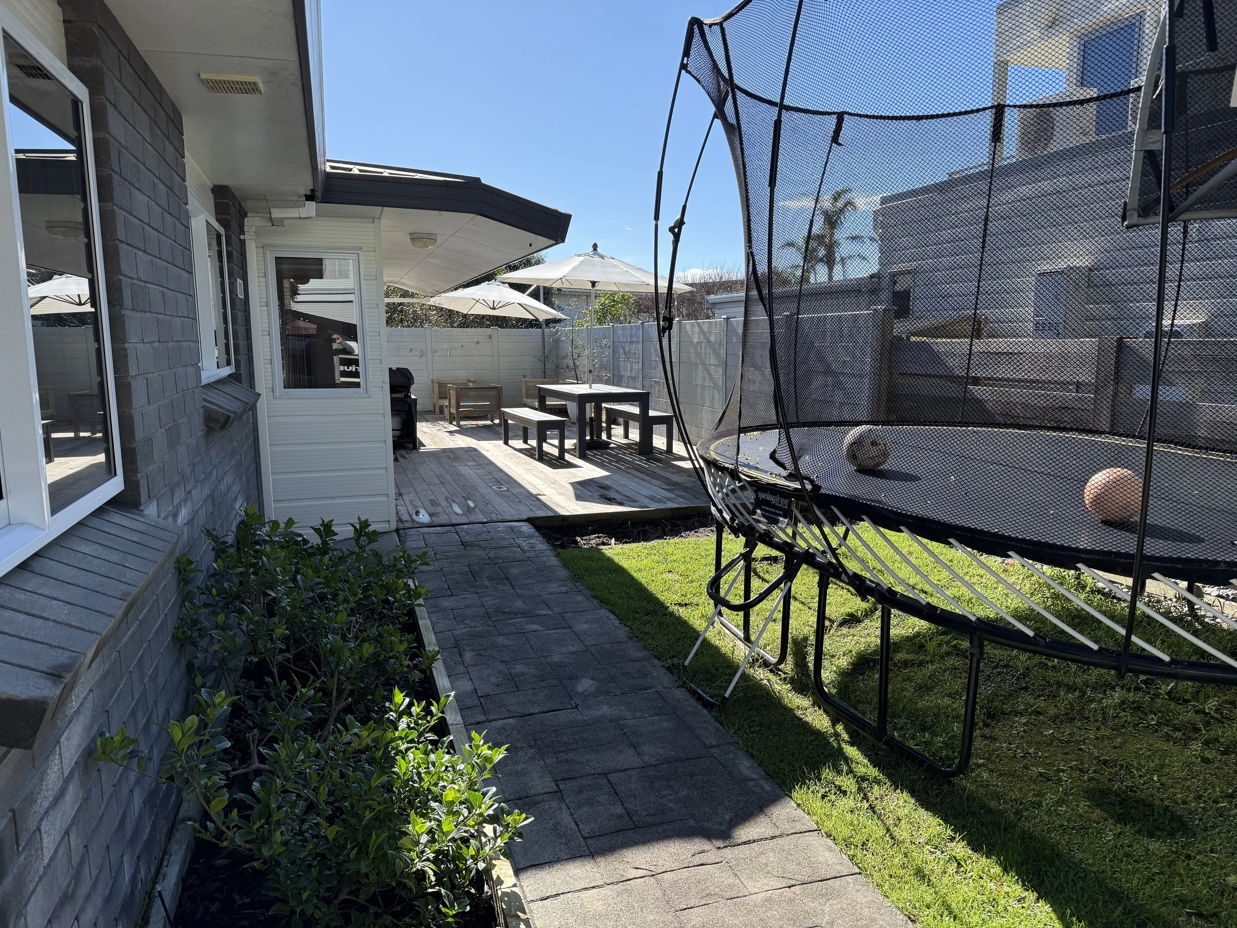 Backyard with patio, outdoor furniture, umbrellas, trampoline with balls, garden, and neighboring houses under a sunny sky.