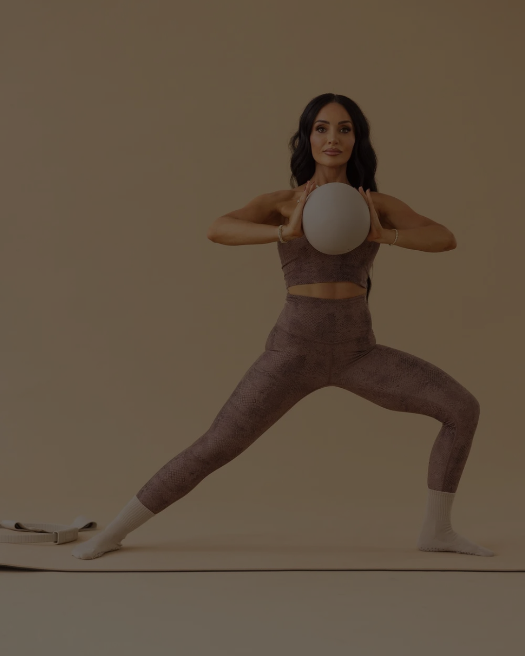 A Pilates instructor demonstrating a grounded mat exercise while holding a stability ball, emphasizing balance, strength, and controlled movement against a minimalist backdrop.