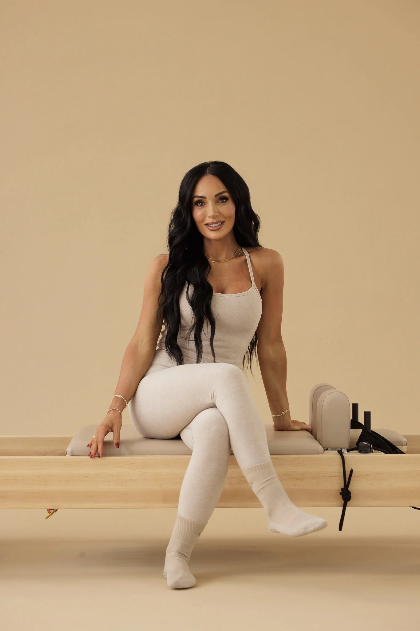 A Pilates instructor sits confidently on a reformer against a warm neutral backdrop, wearing light-toned activewear, with long dark hair styled loose and a calm, welcoming expression in a minimalist studio setting.